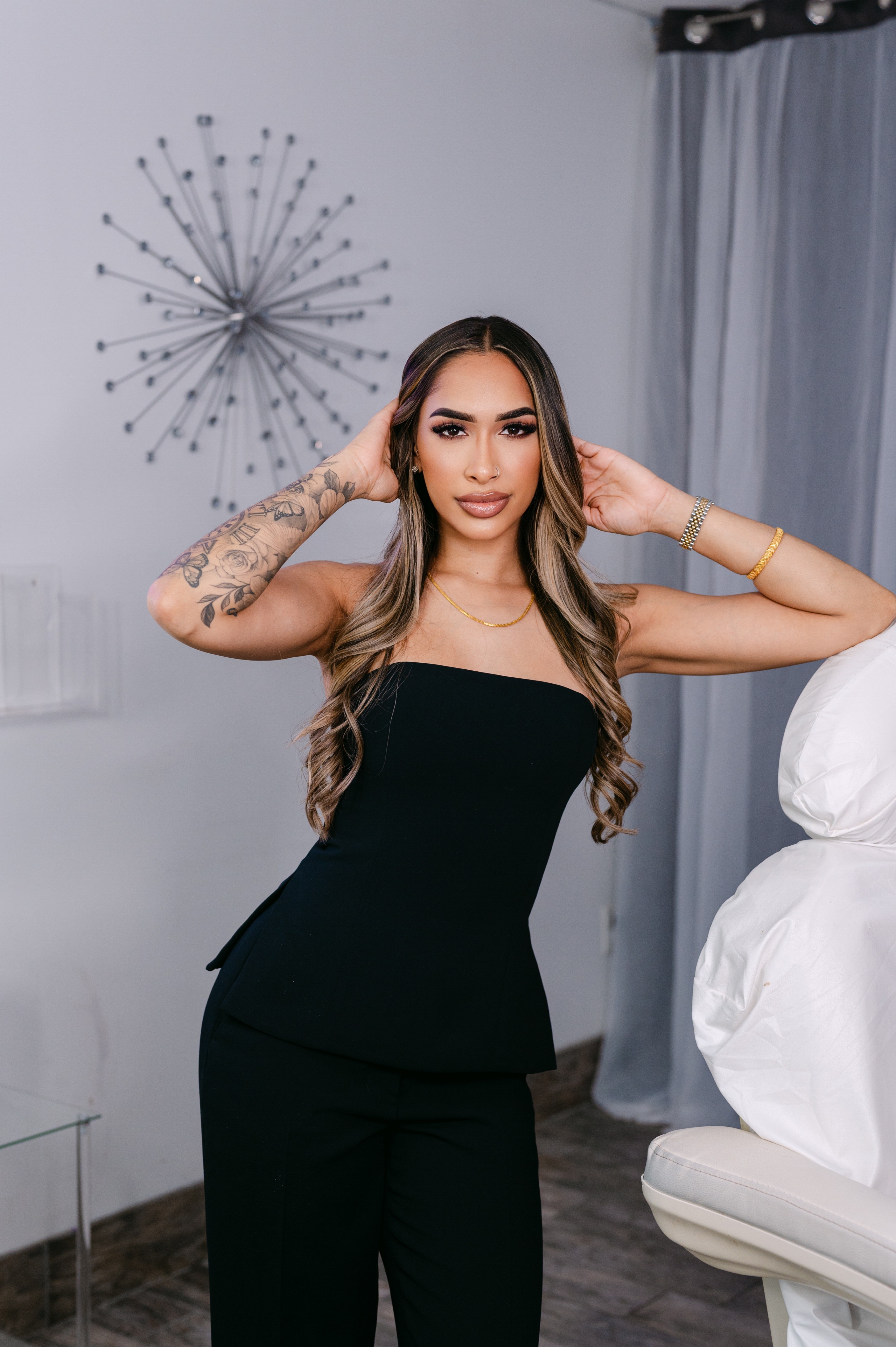 Woman with long hair and tattoos wearing a black strapless outfit, posing indoors with a decorative wall piece behind her.
