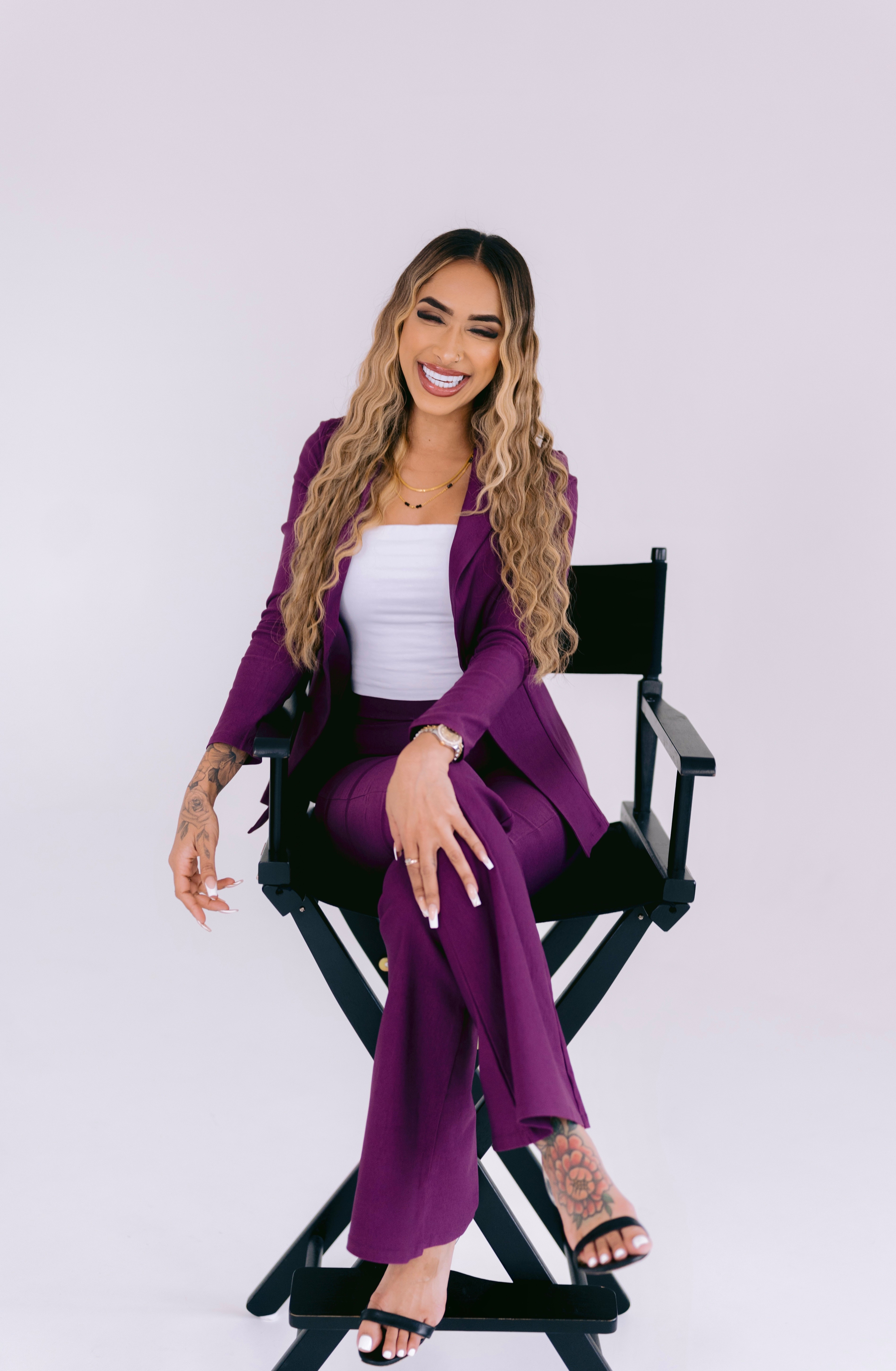 Woman with long wavy hair smiling, sitting on a black director's chair, wearing a purple suit and white top.