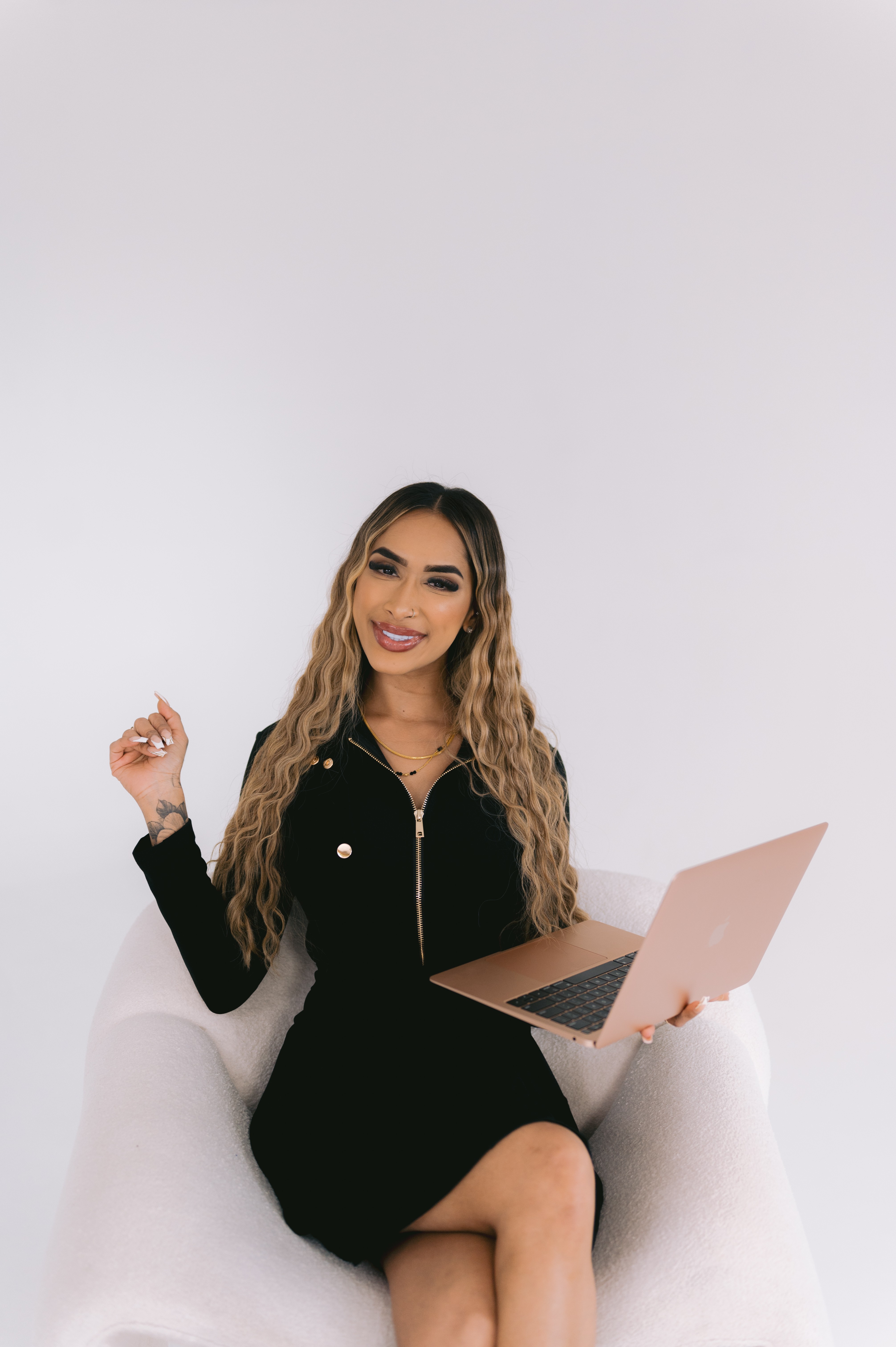 Woman with long wavy hair sitting on a white chair, holding a laptop, smiling, against a plain white background.