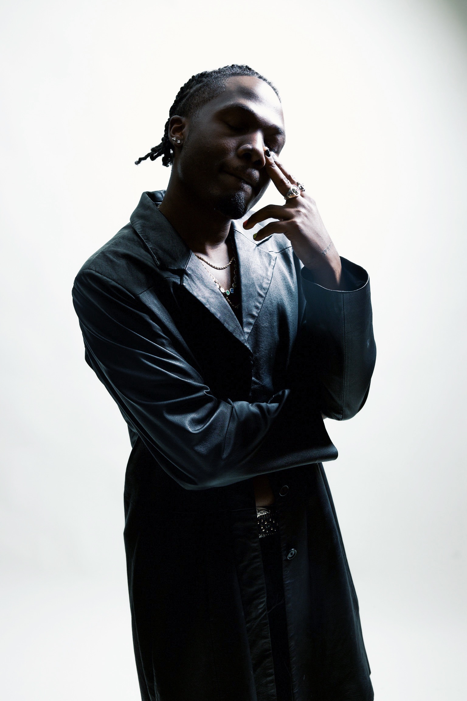 Person with braided hair touching face, wearing dark jacket, standing against a plain light background.