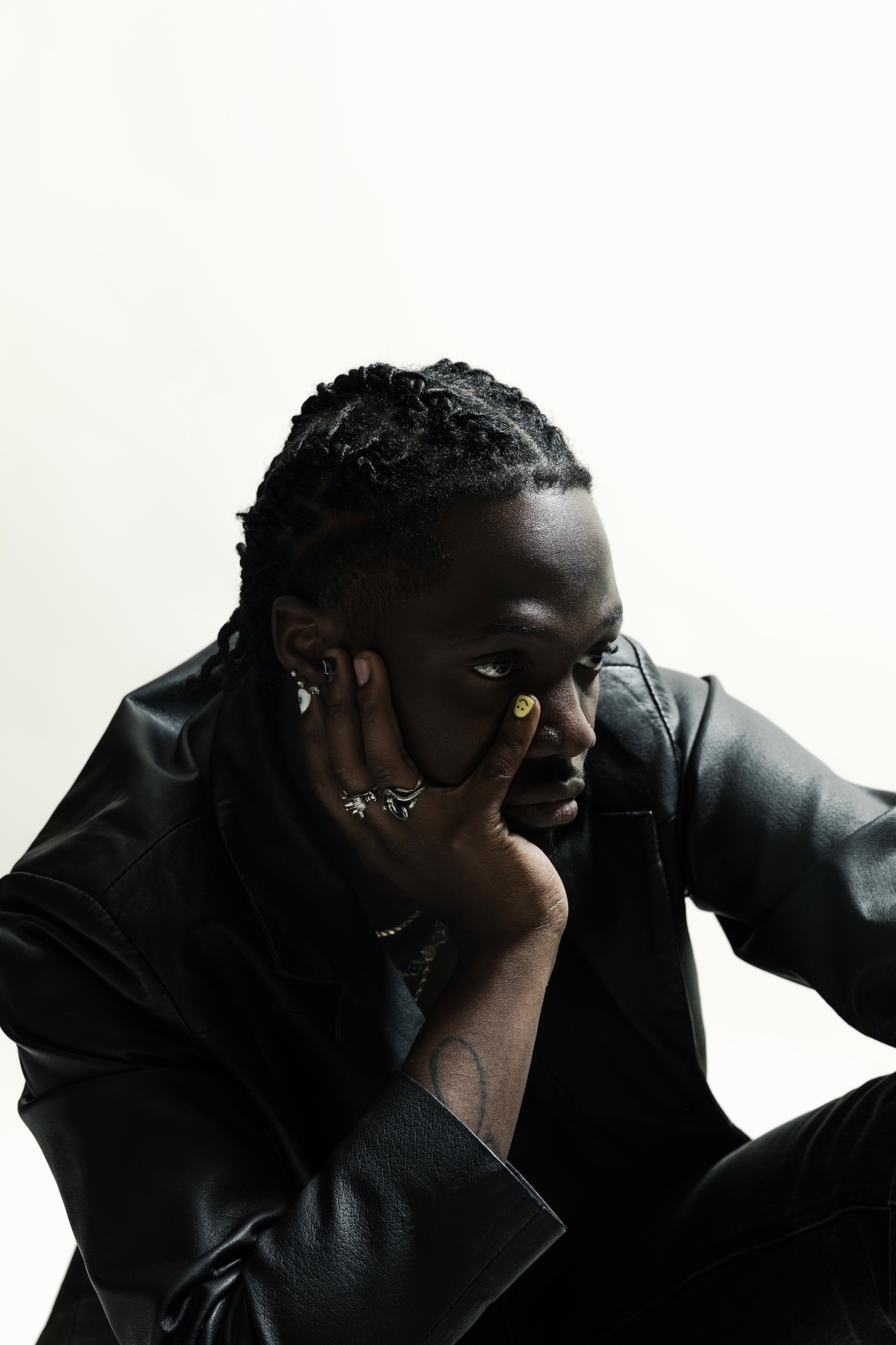 Person with braided hair resting their face on their hand, wearing a black leather jacket, against a plain white background.