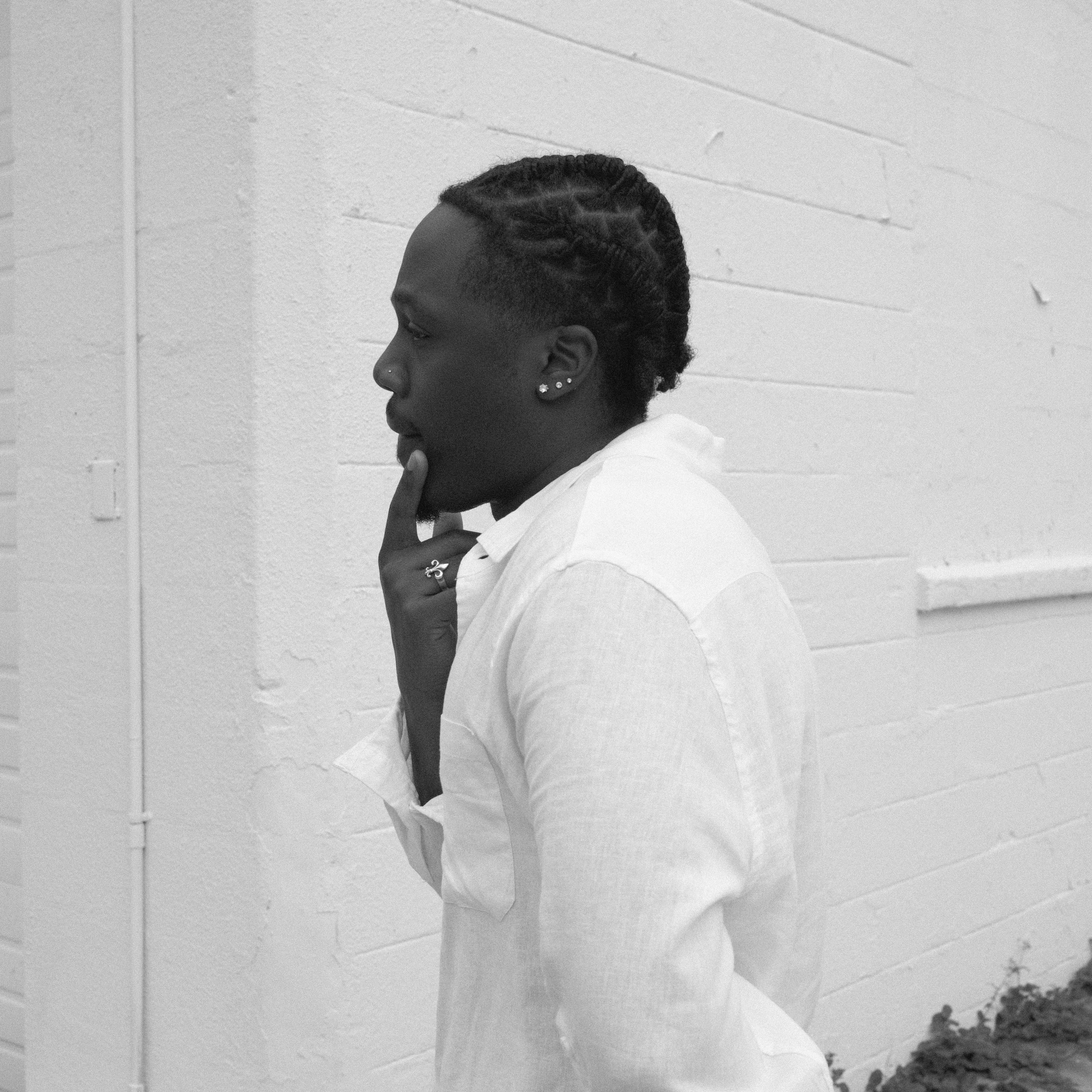 Person with braided hair in profile, wearing a white shirt, standing against a brick wall, touching chin thoughtfully.