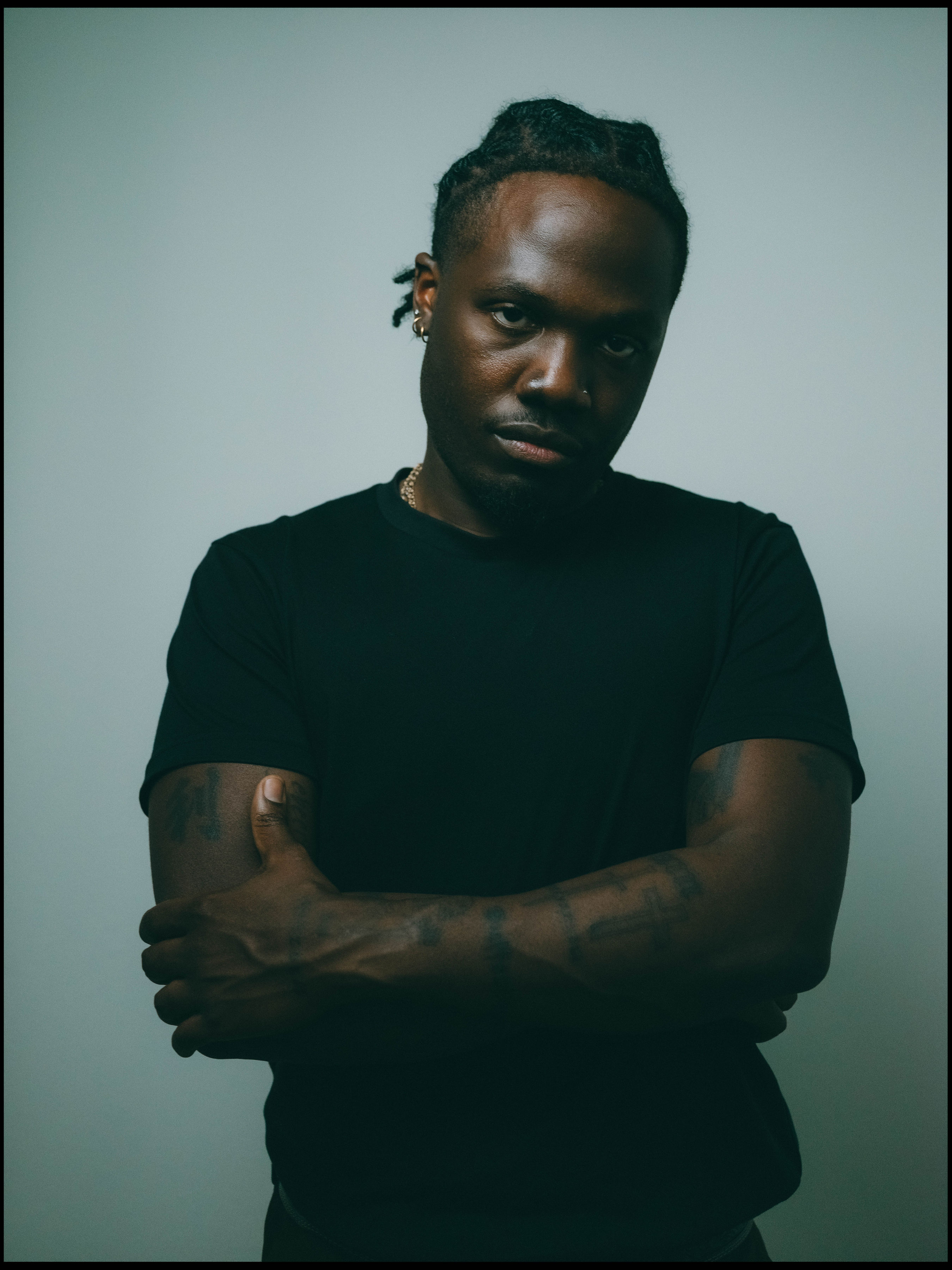 Person with crossed arms, wearing a black shirt, looking at the camera against a plain background.
