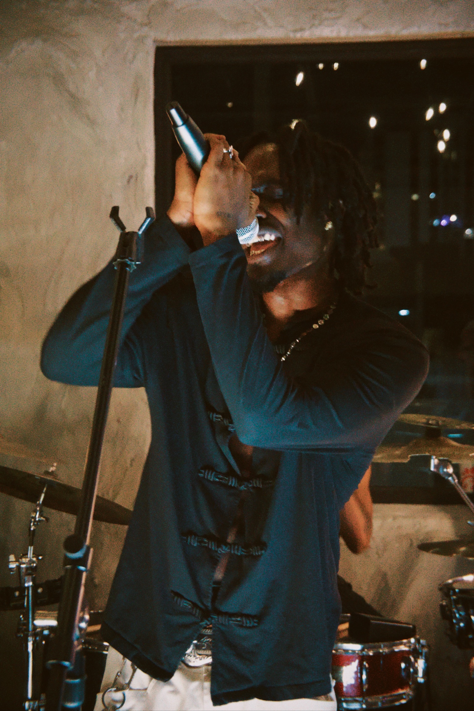 Person singing into a microphone with a smile, wearing a dark jacket, in a dimly lit setting.