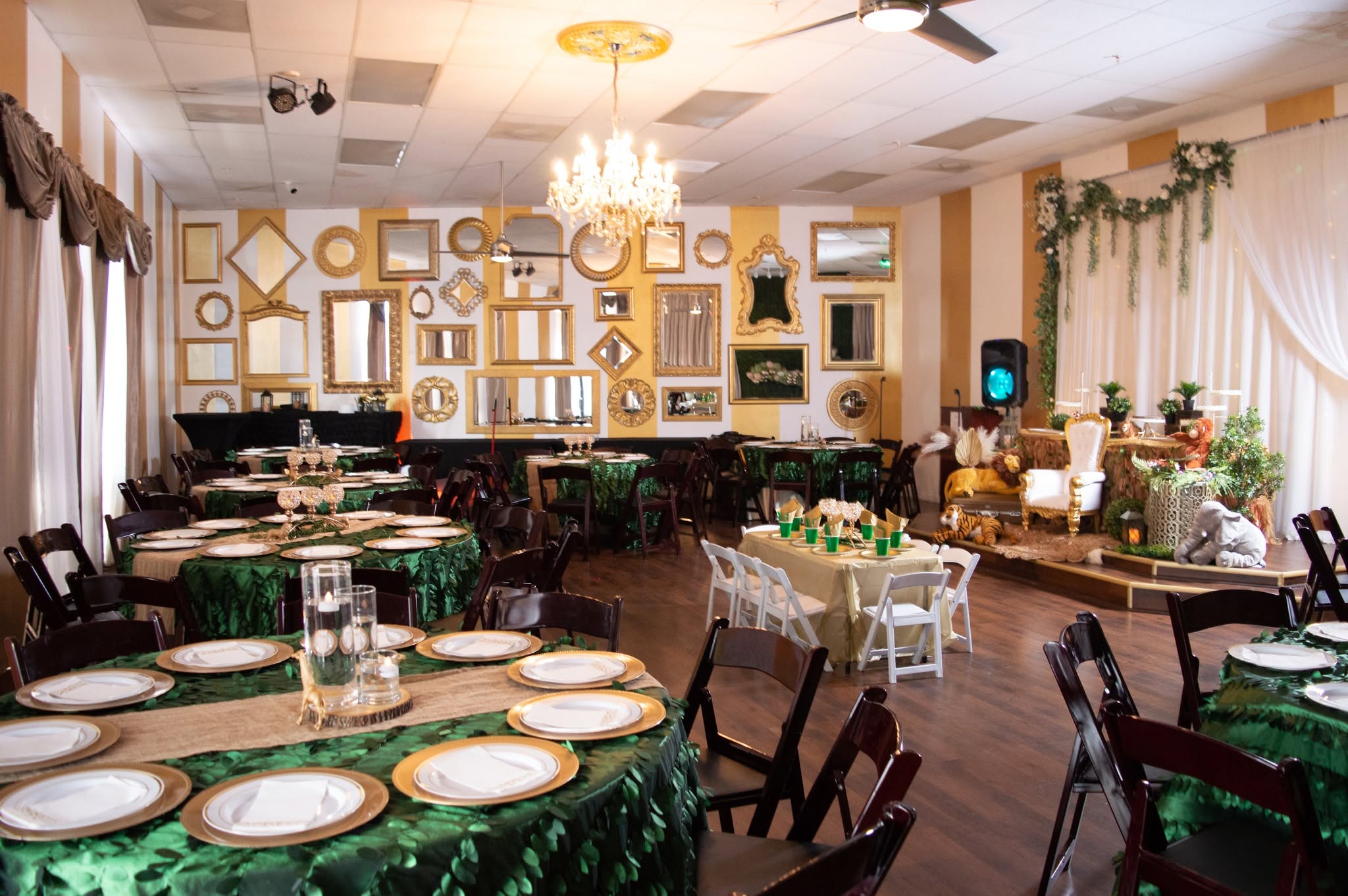 Elegant event room with round tables covered in green tablecloths, set with plates and glasses, and decorated with a chandelier and greenery.