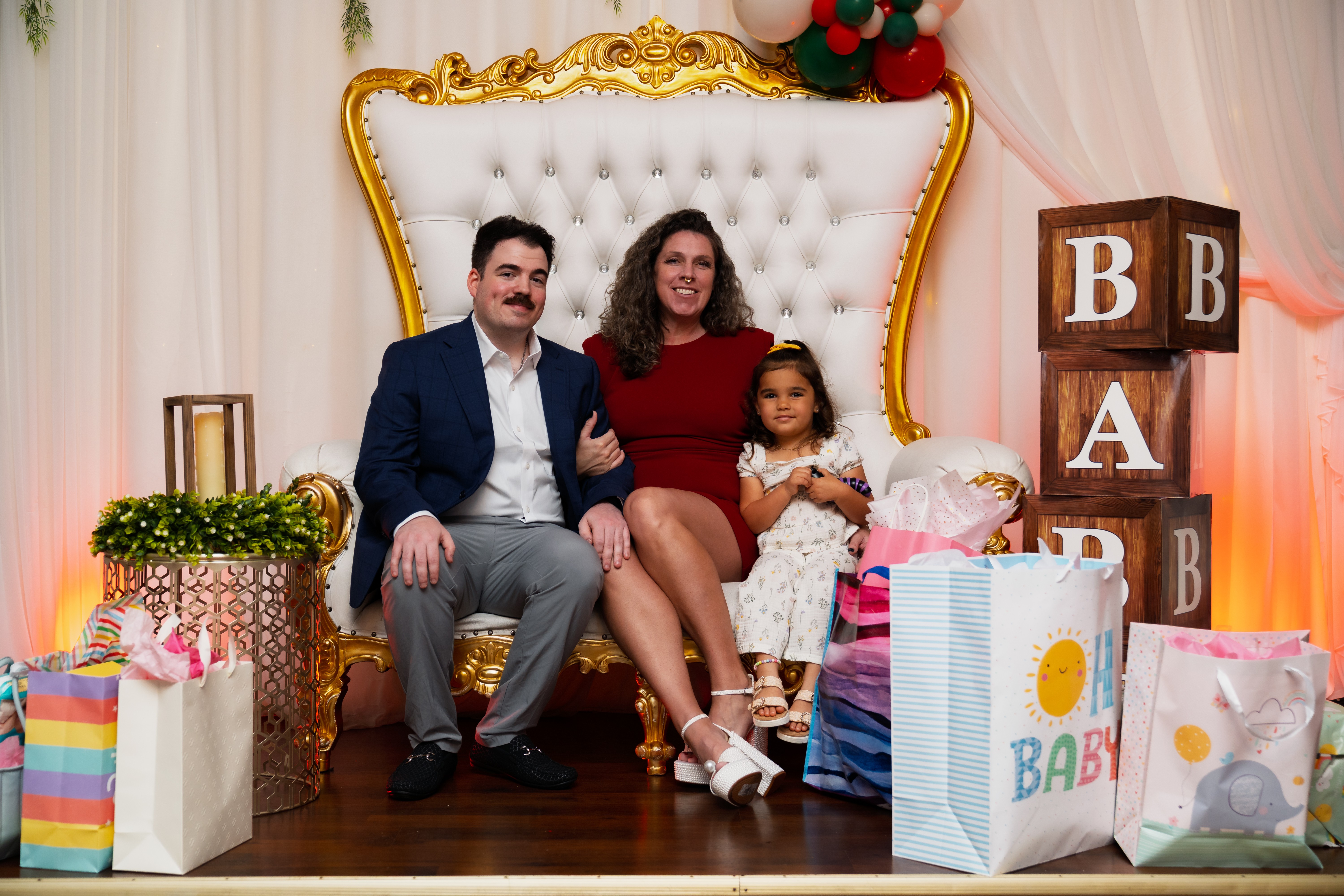 Family sitting on a decorated throne-like chair with gifts around, celebrating a special occasion, with a woman, man, and girl.