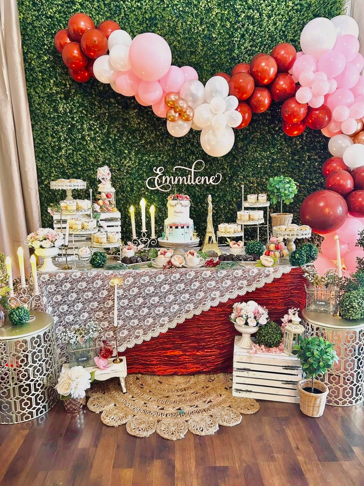 Decorated birthday table with balloons, cake, and floral arrangements, with a green hedge backdrop and wooden floor.