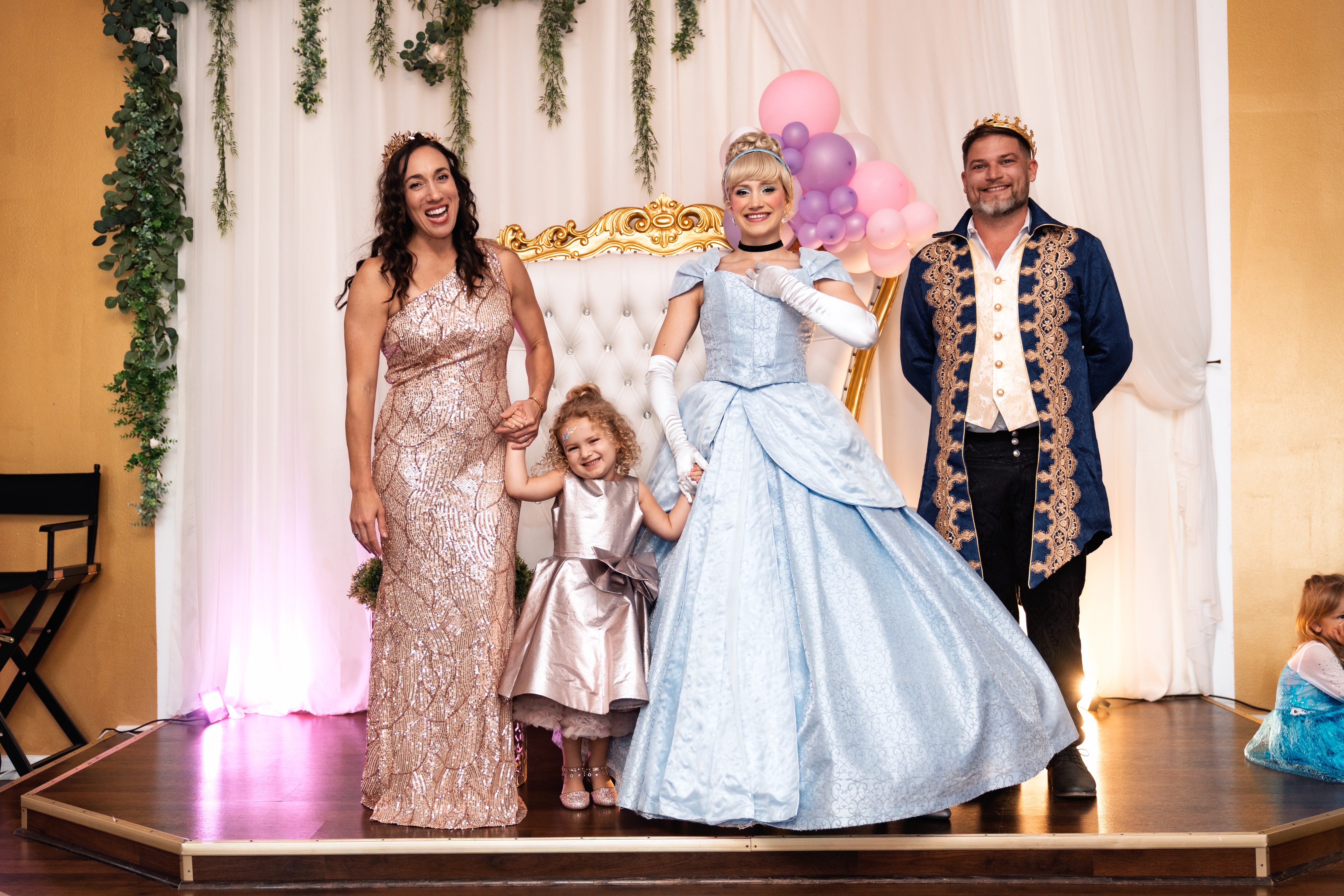 Four people on stage, two women, a girl, and a man, with a decorated backdrop and balloons, smiling and posing.