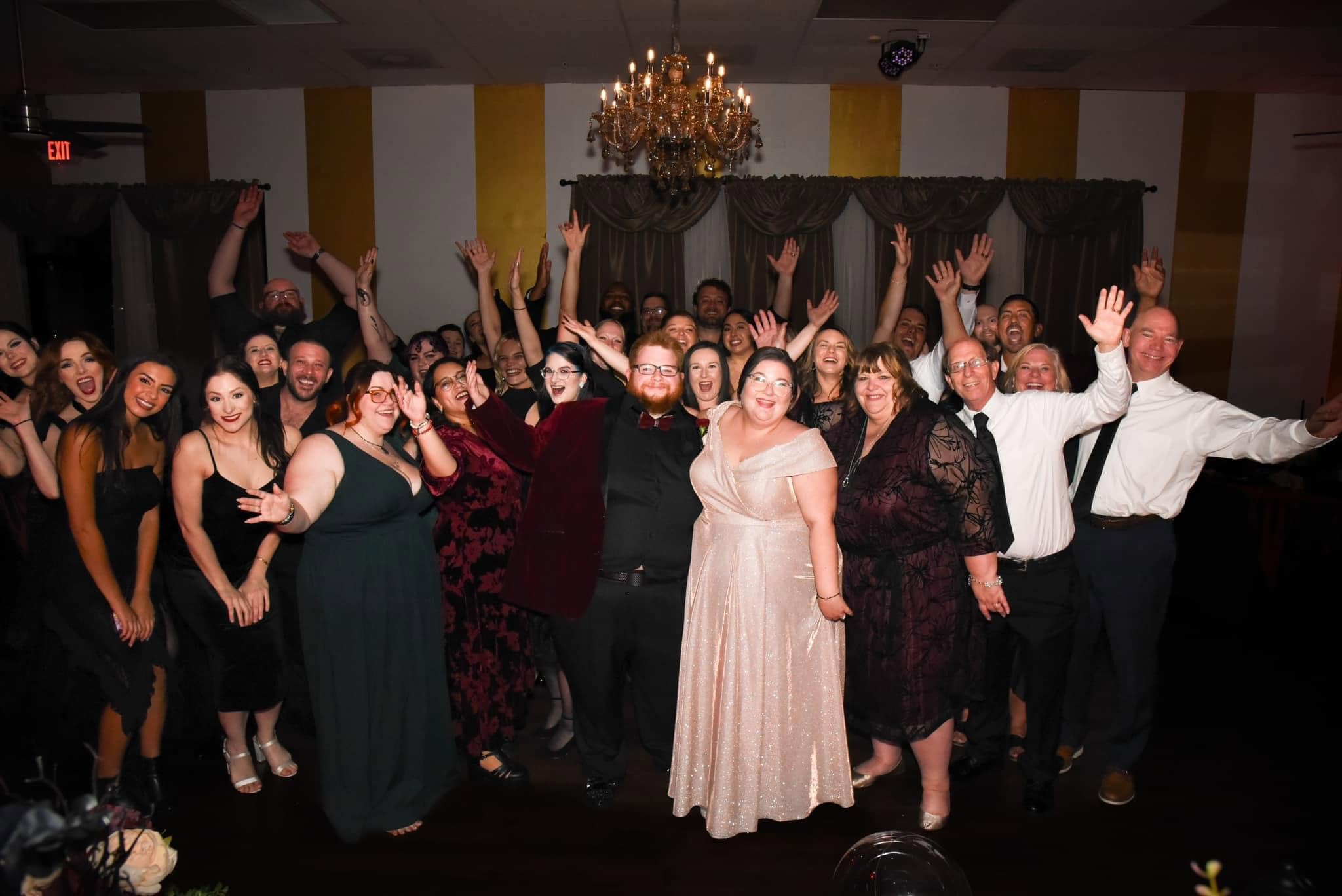 Group of people celebrating indoors, some with arms raised, dressed in formal attire, with a chandelier overhead.
