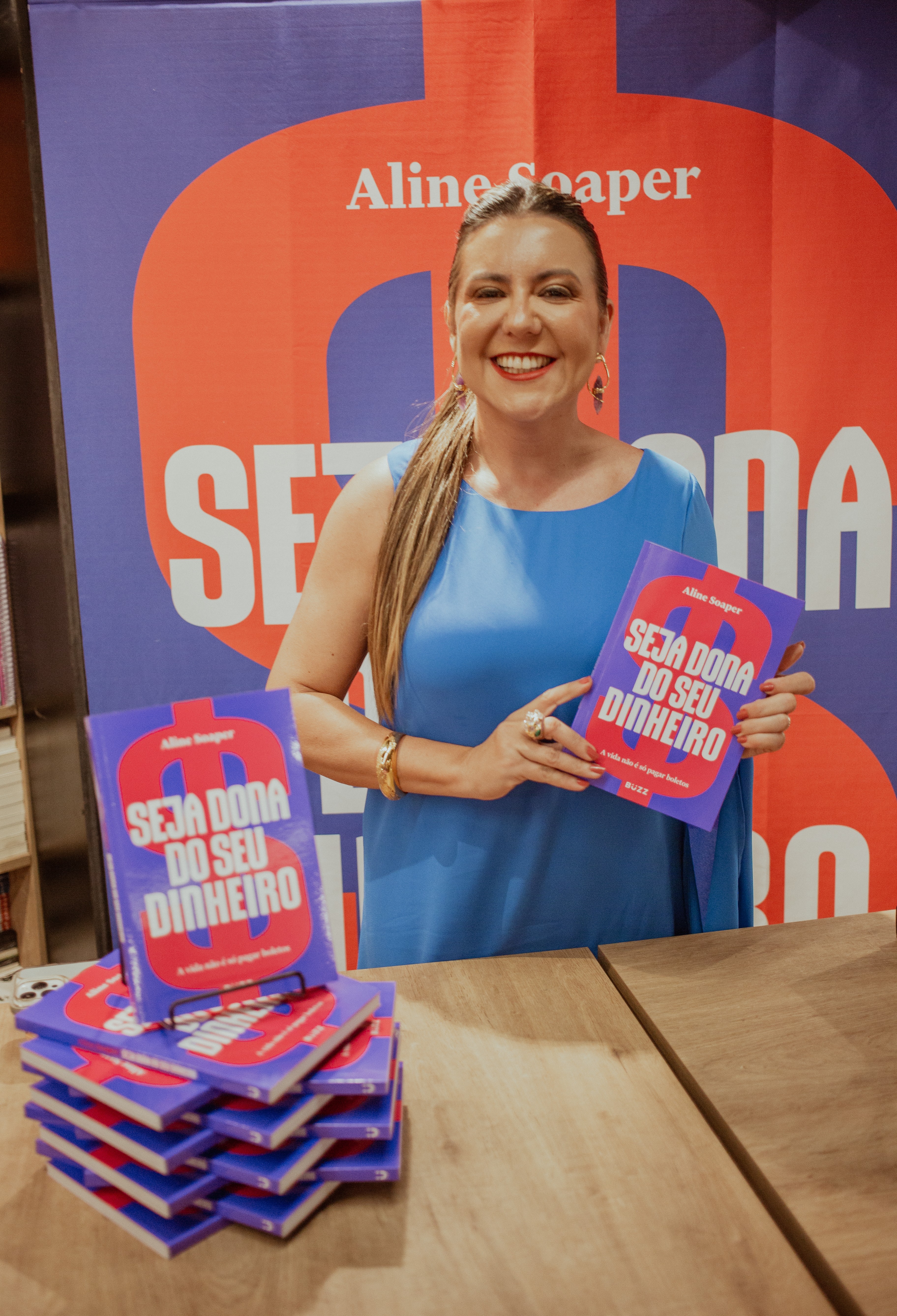 Smiling woman in blue dress holding a book, standing in front of a promotional backdrop, with a stack of books on a table.