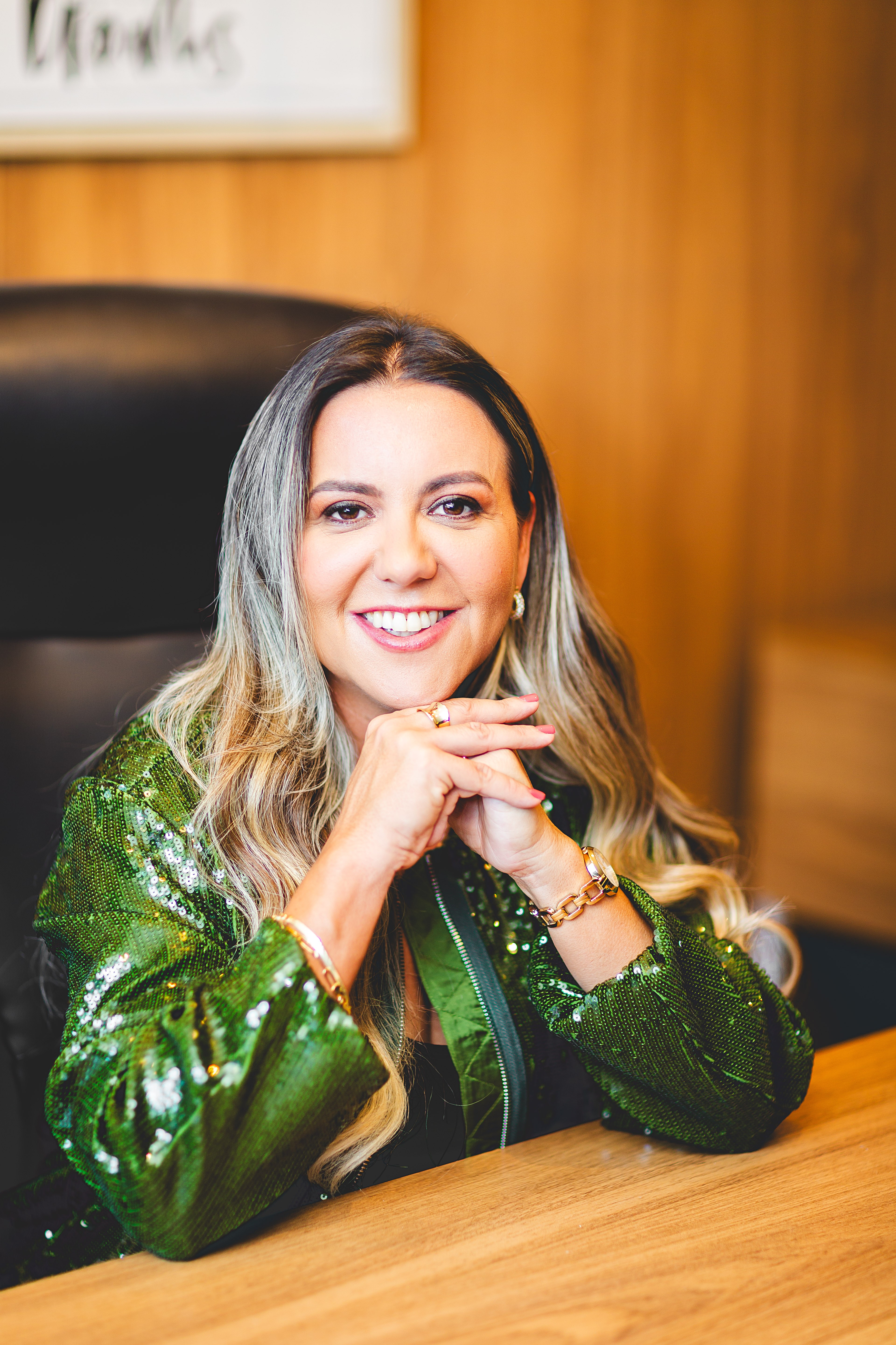 Woman with long wavy hair smiling, wearing a shiny green jacket, sitting at a wooden table, with a wooden wall background.