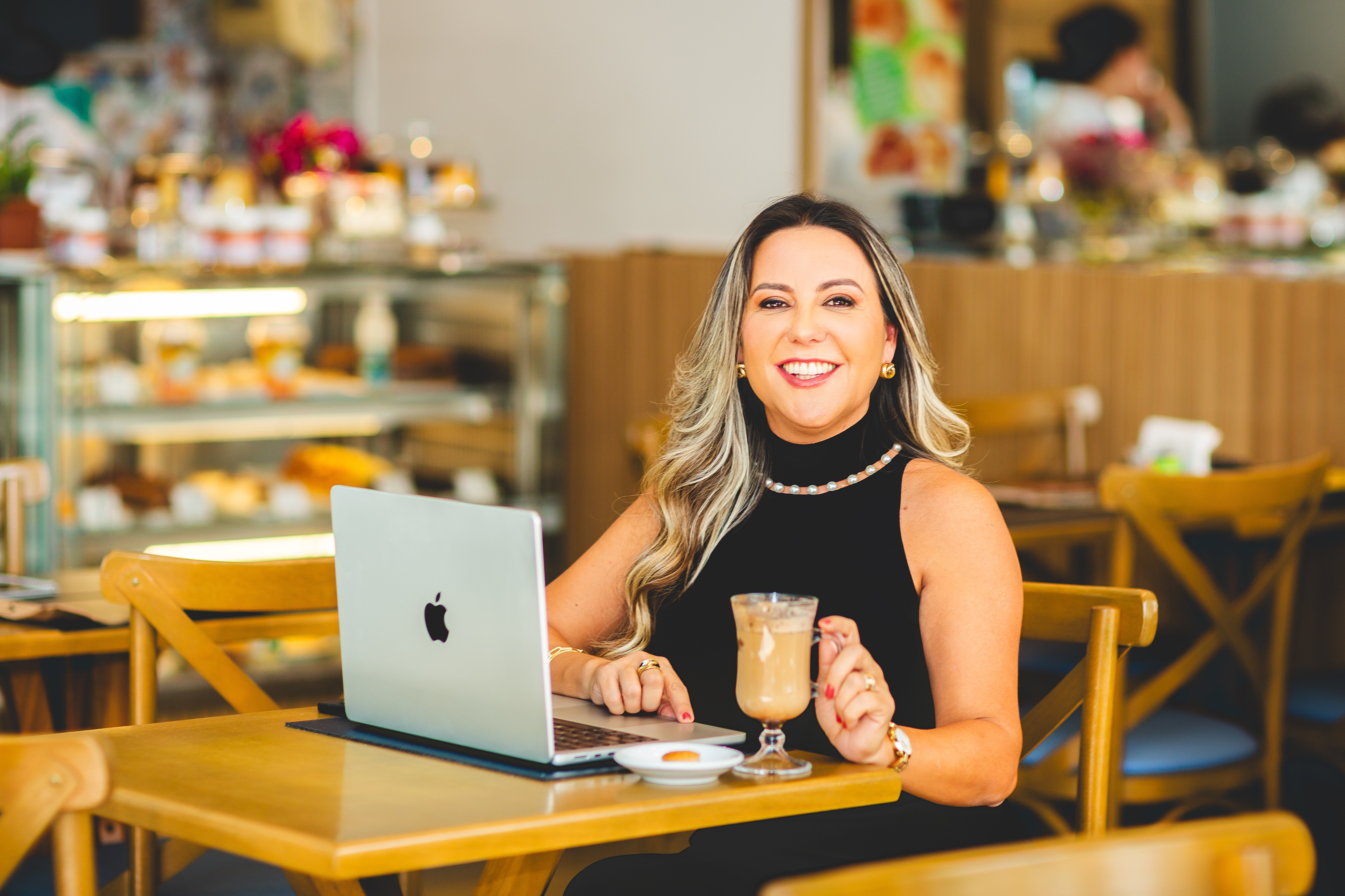 Woman with long hair smiling at a laptop in a cafe, holding a drink, with a blurred background of baked goods.