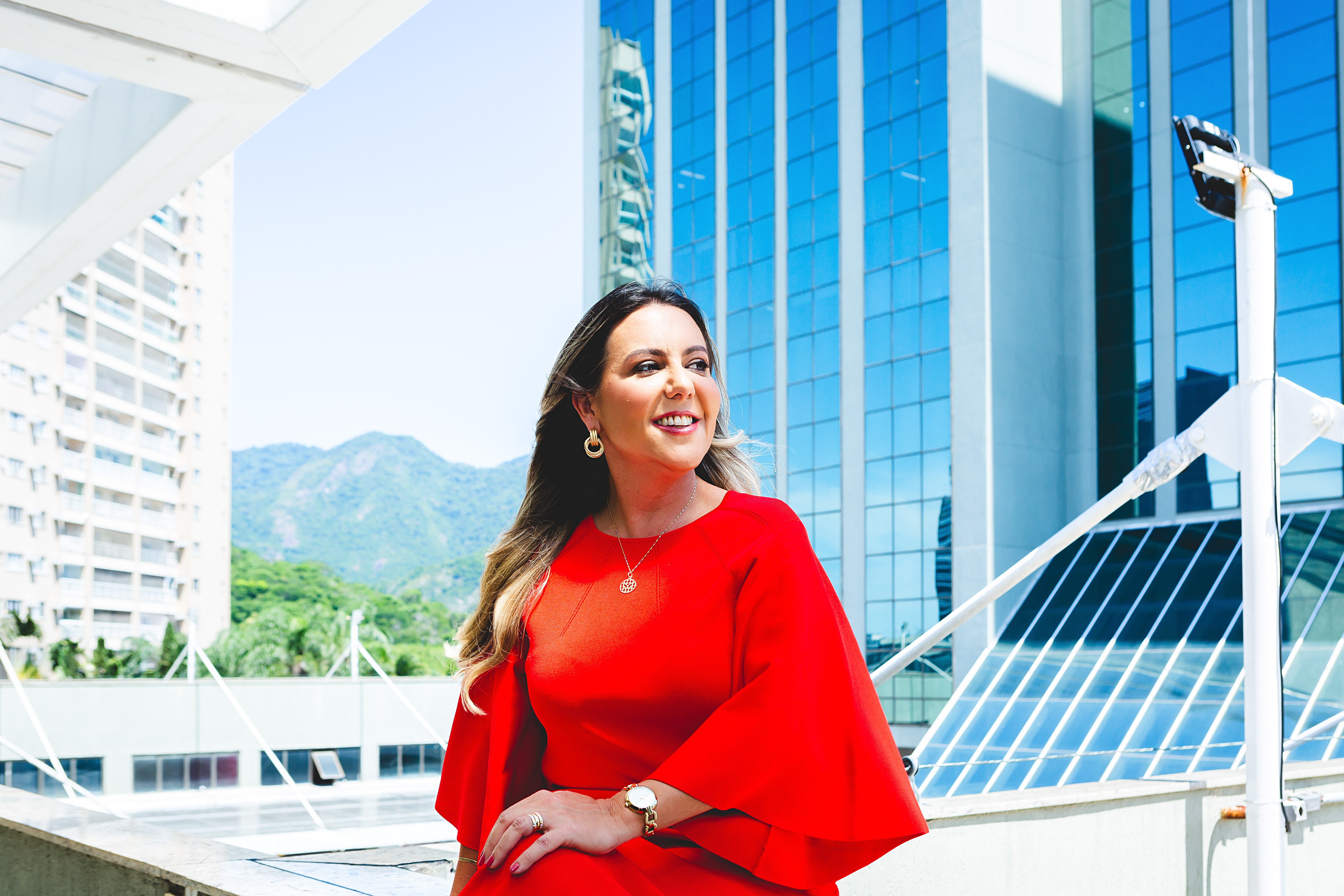 Woman in red dress smiling outdoors with modern glass buildings and mountains in background.