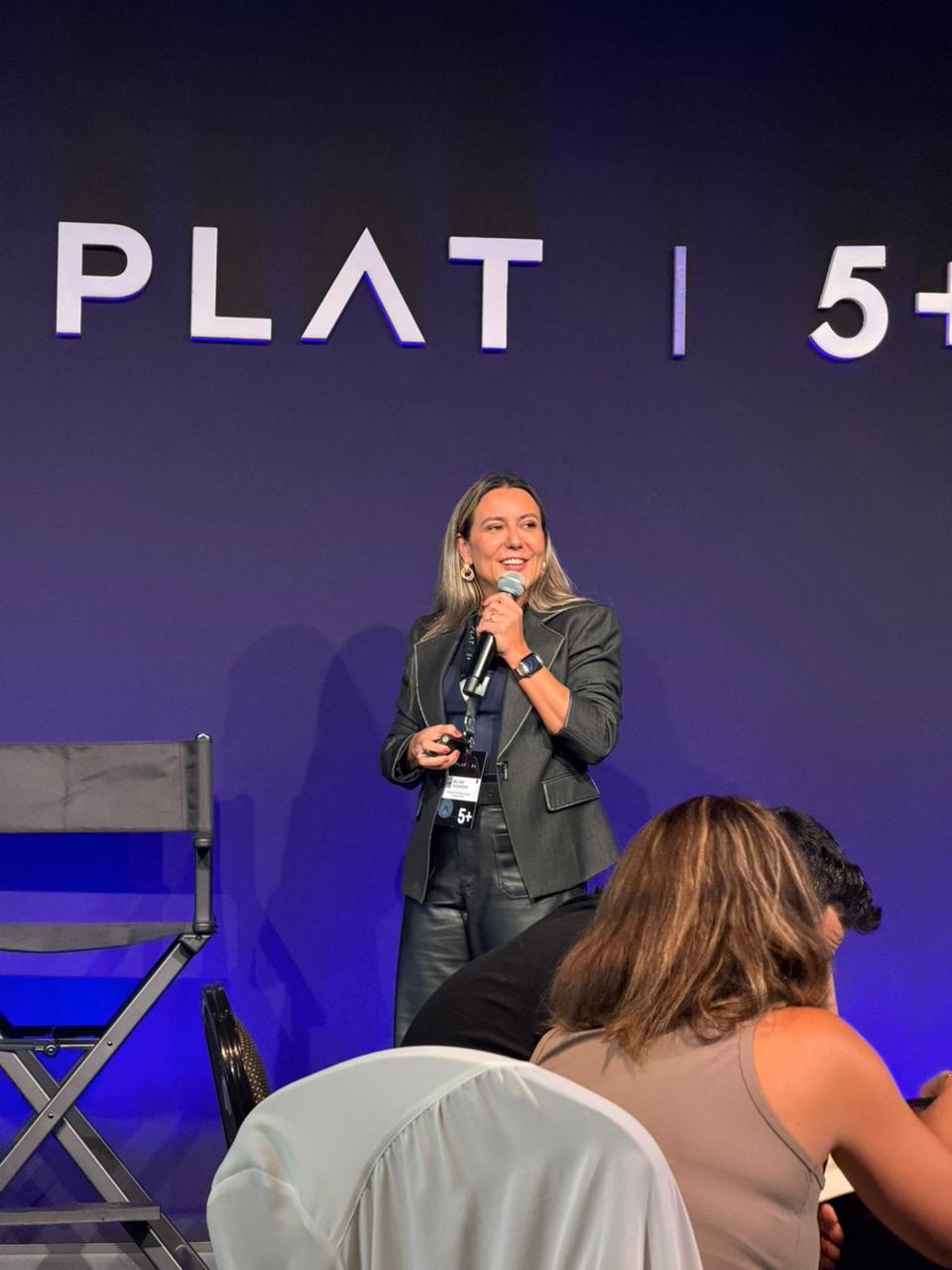 Woman speaking on stage with a microphone, standing in front of a dark background with text, audience members in foreground.