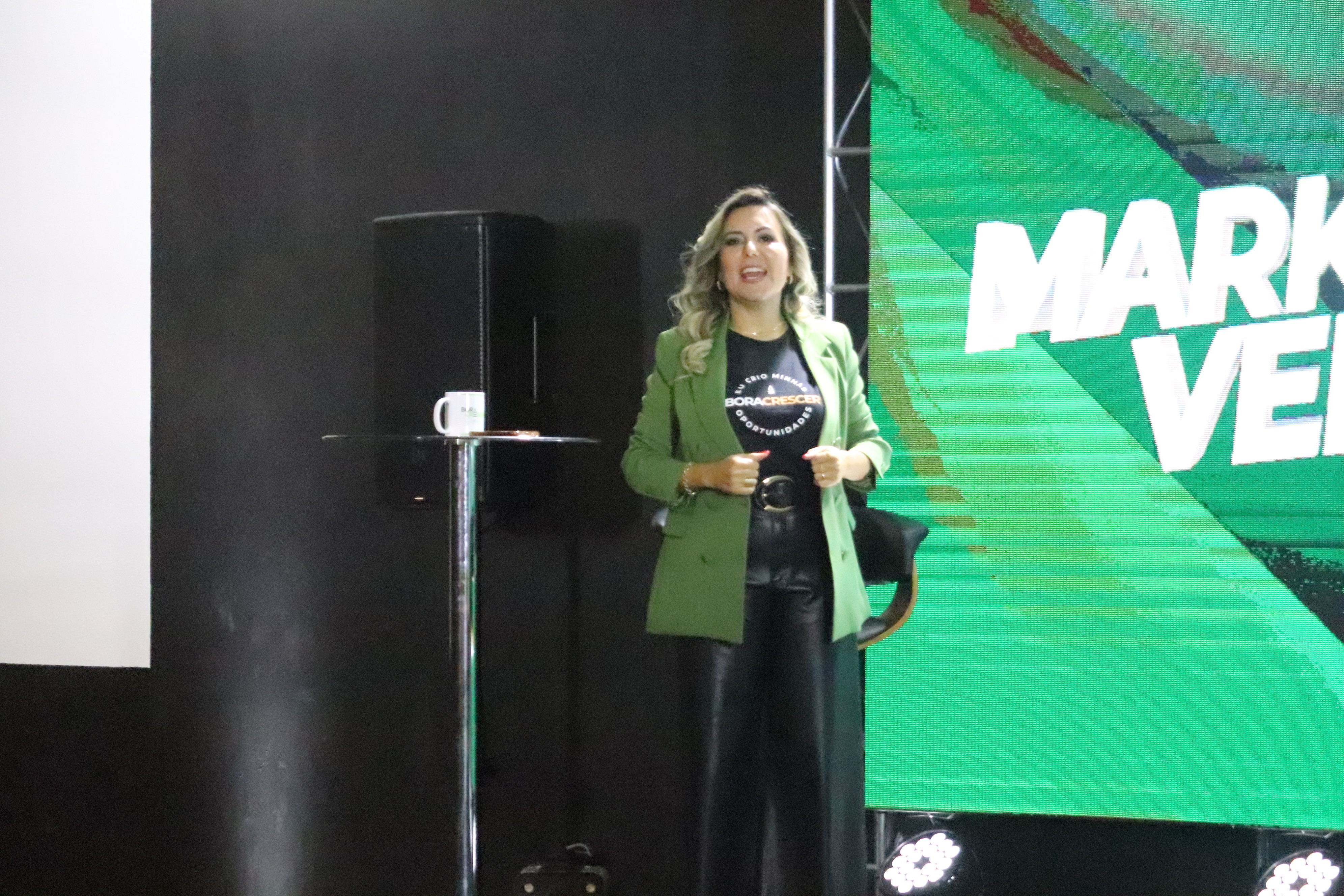 Woman standing on stage with a large green screen behind her, holding a microphone, smiling, wearing a green blazer.