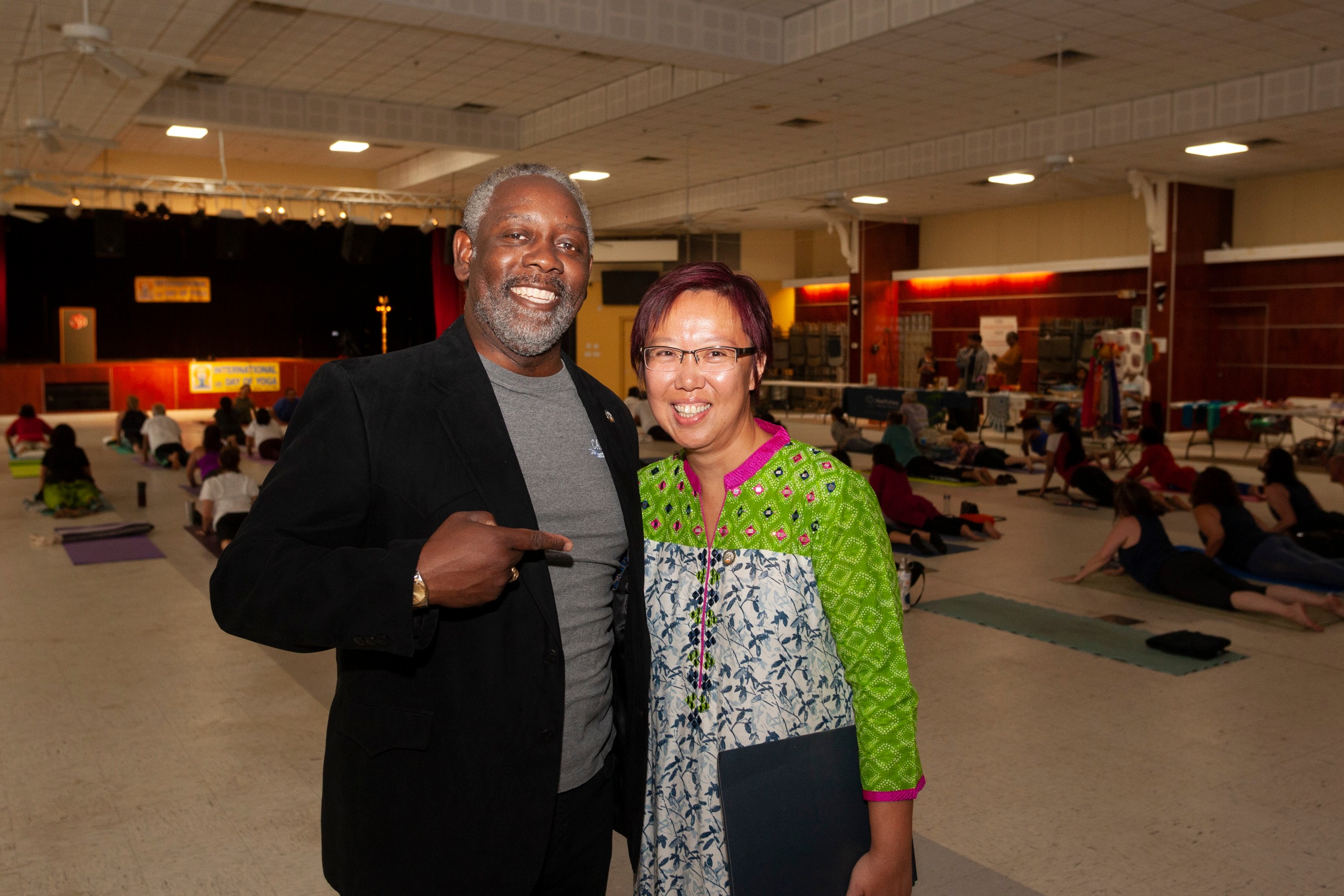 Two people smiling indoors, one pointing at the other, with a group practicing yoga in the background.