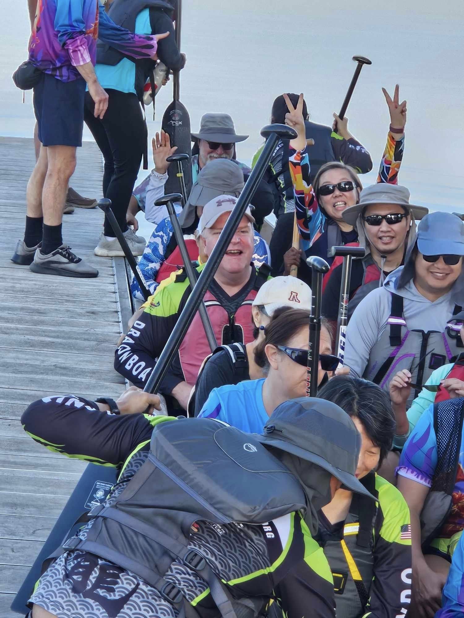 Group of people in life jackets and hats on a dock, preparing for kayaking or boating, some making peace signs.