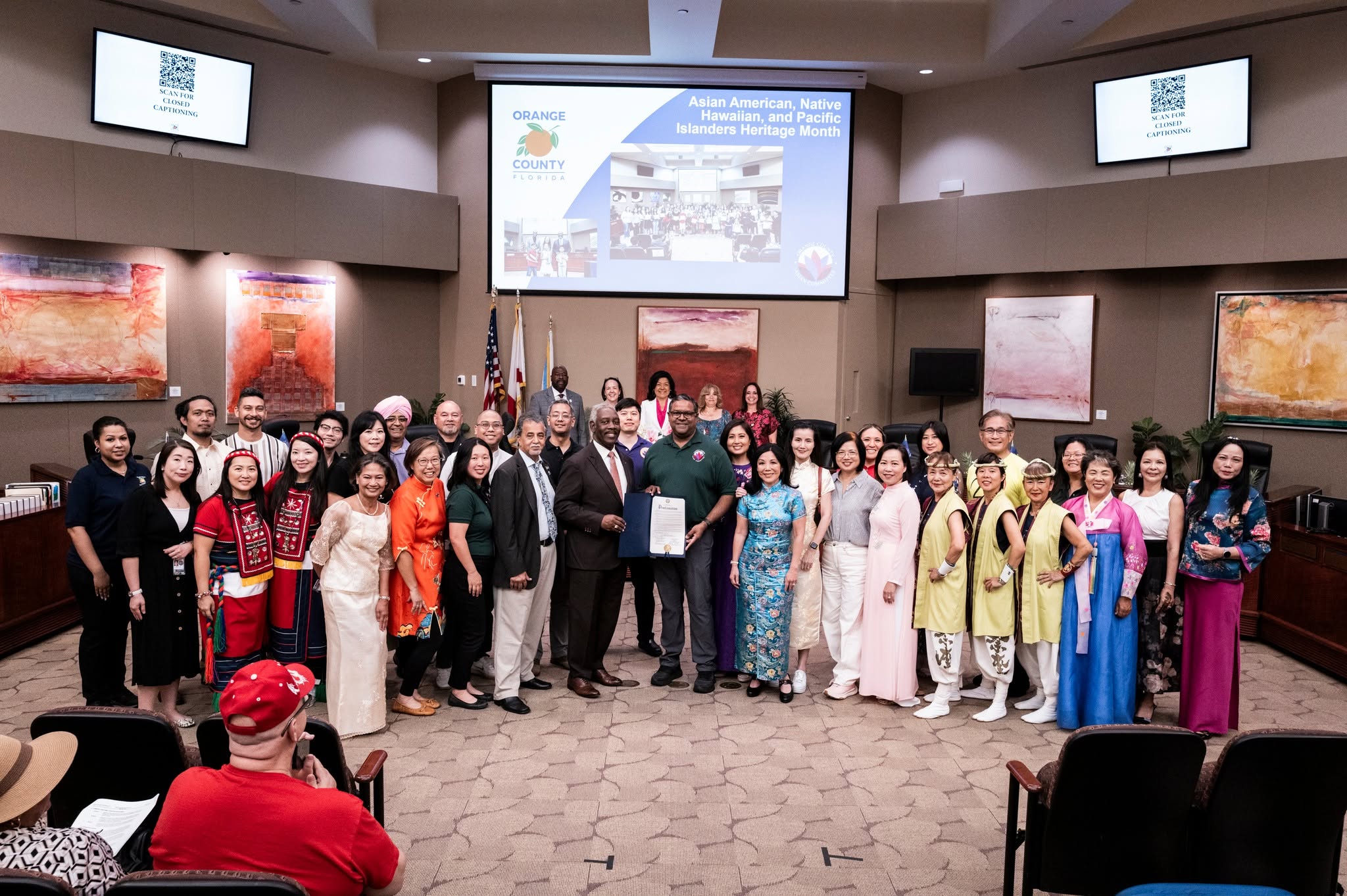 Group of people standing in a conference room with screens and artwork, some holding a certificate, audience seated in front.