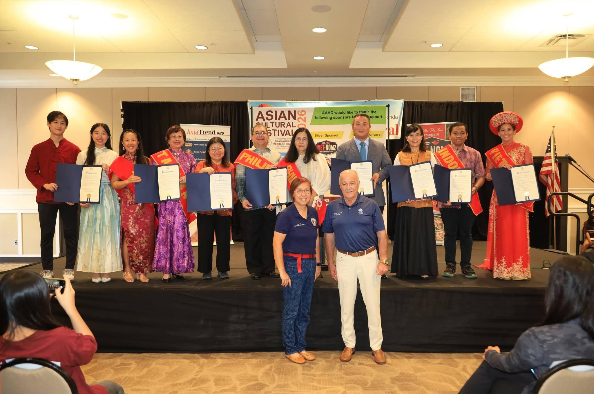 Group of people on stage holding certificates, with two individuals in front, in a conference room setting.