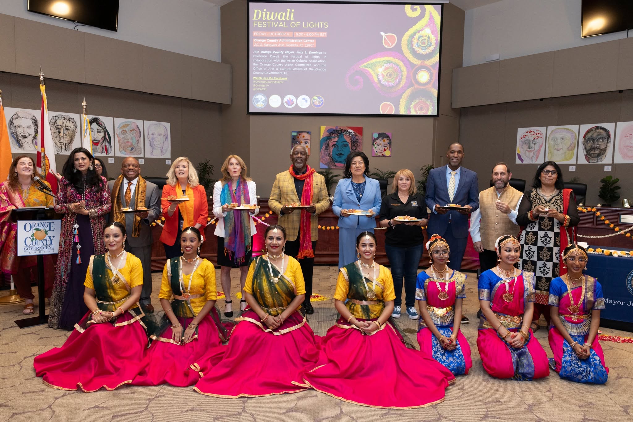 Group of people on stage, some in colorful traditional attire, with artwork displayed on walls behind them.