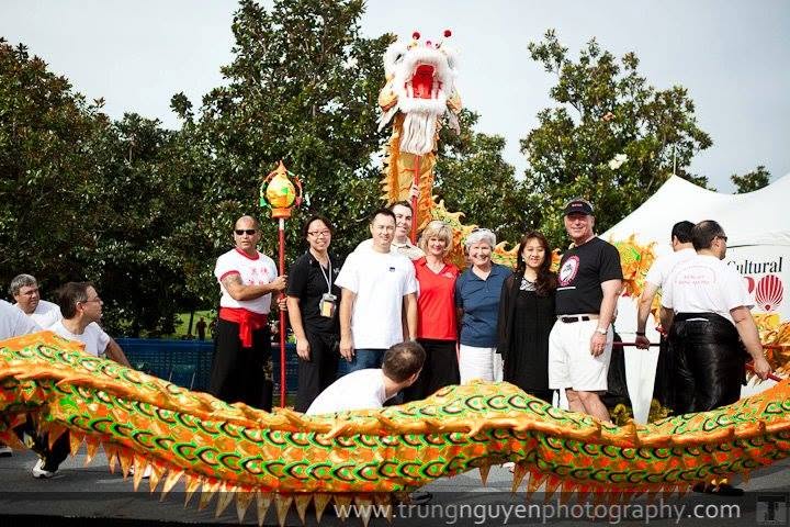 Group of people standing outdoors with a dragon costume in front, trees and a tent in the background.