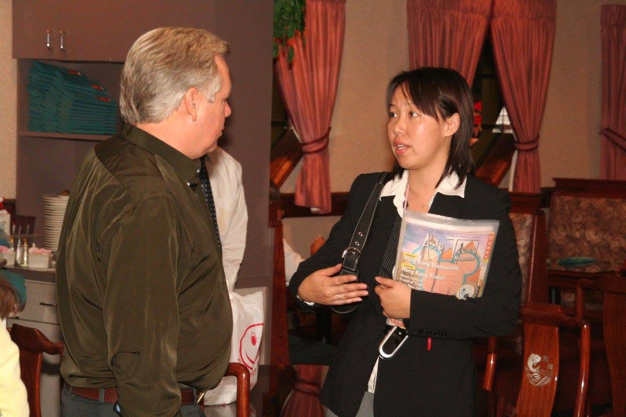 Two people engaged in conversation indoors, with curtains and furniture in the background.