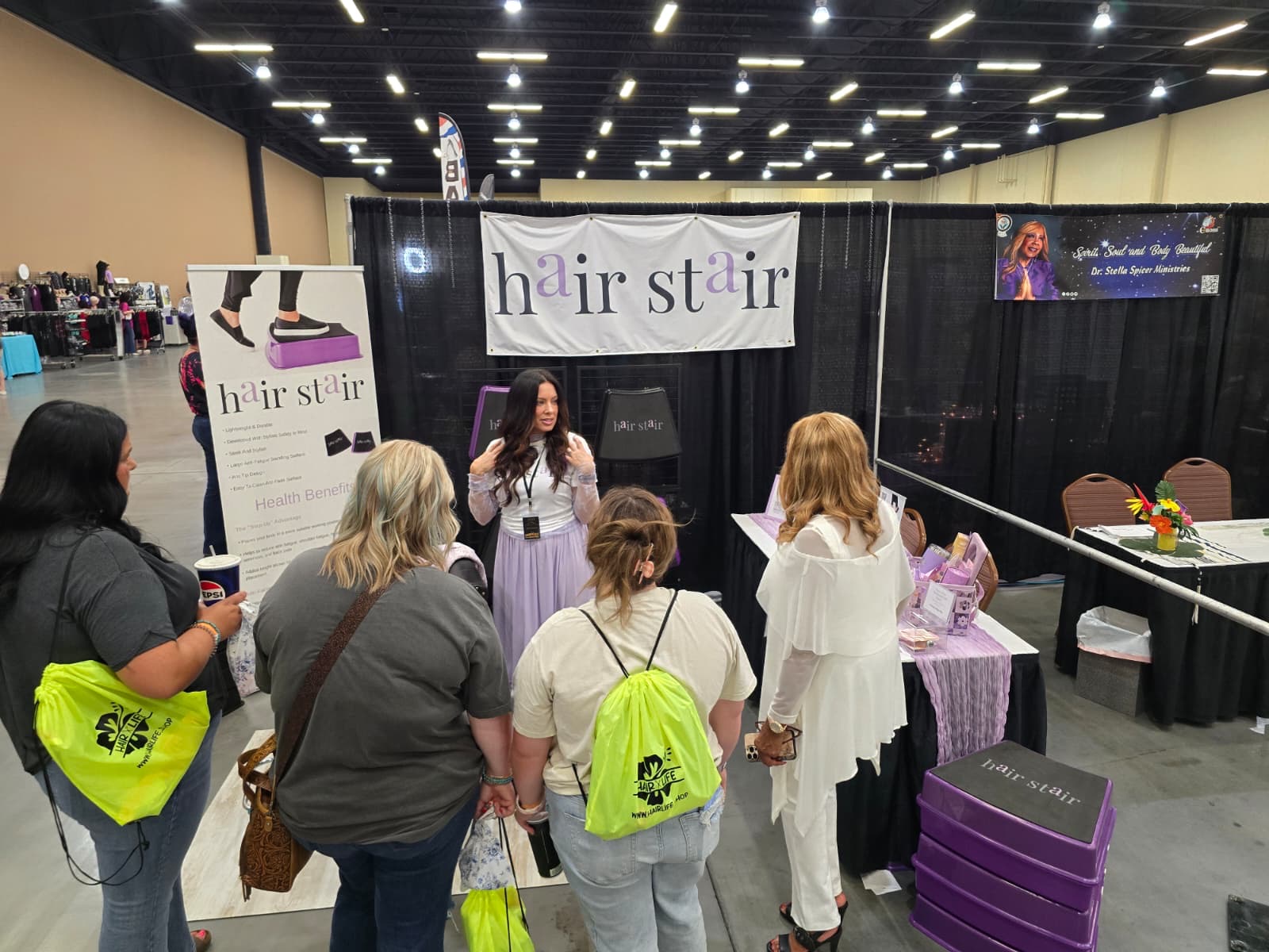Woman speaking to a group at a hair styling booth with a banner reading 'hairstair' behind her, in an indoor event space.