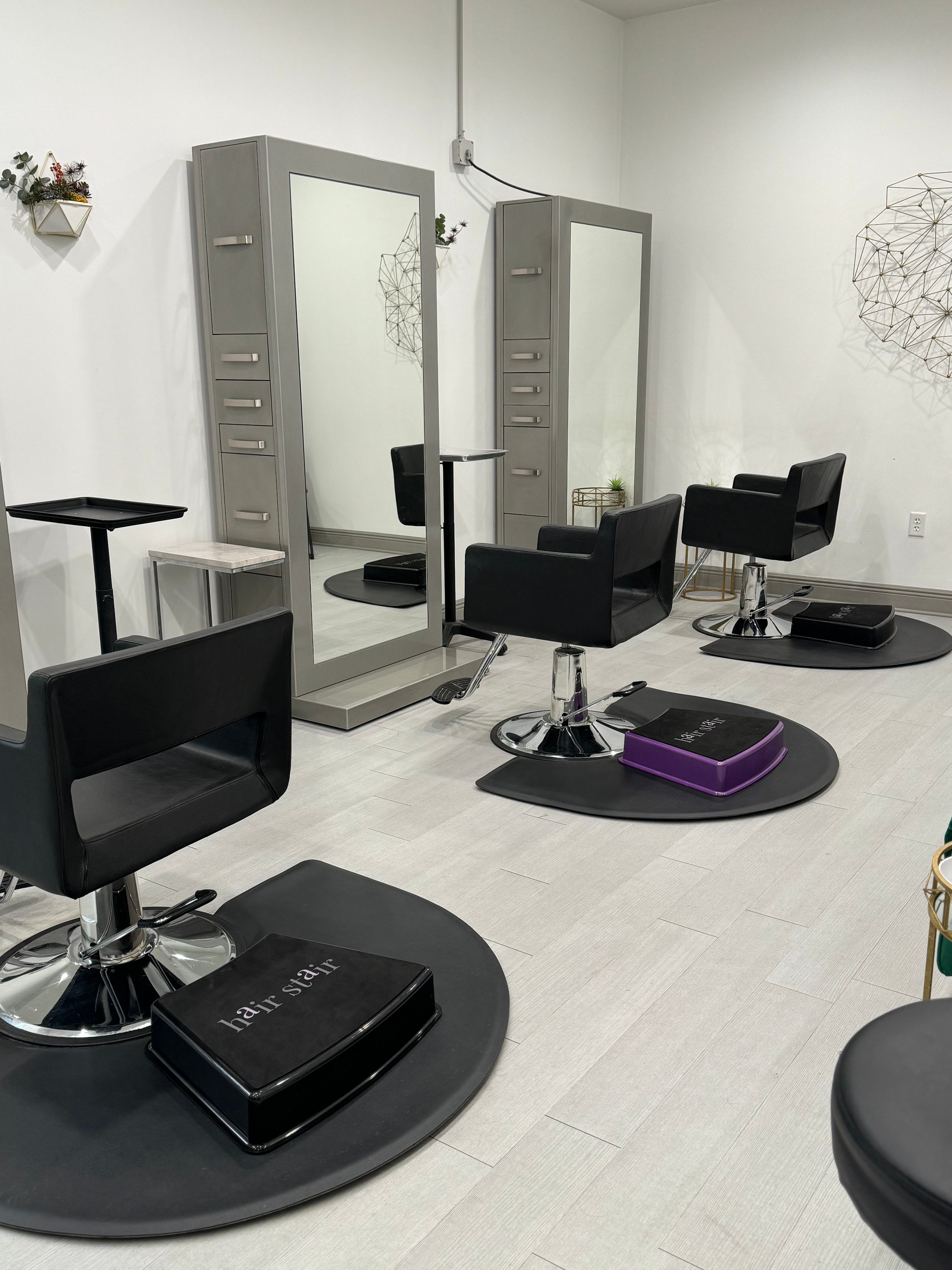 Hair salon with chairs, mirrors, and styling stations, featuring black styling chairs and footrests, and a white wall background.