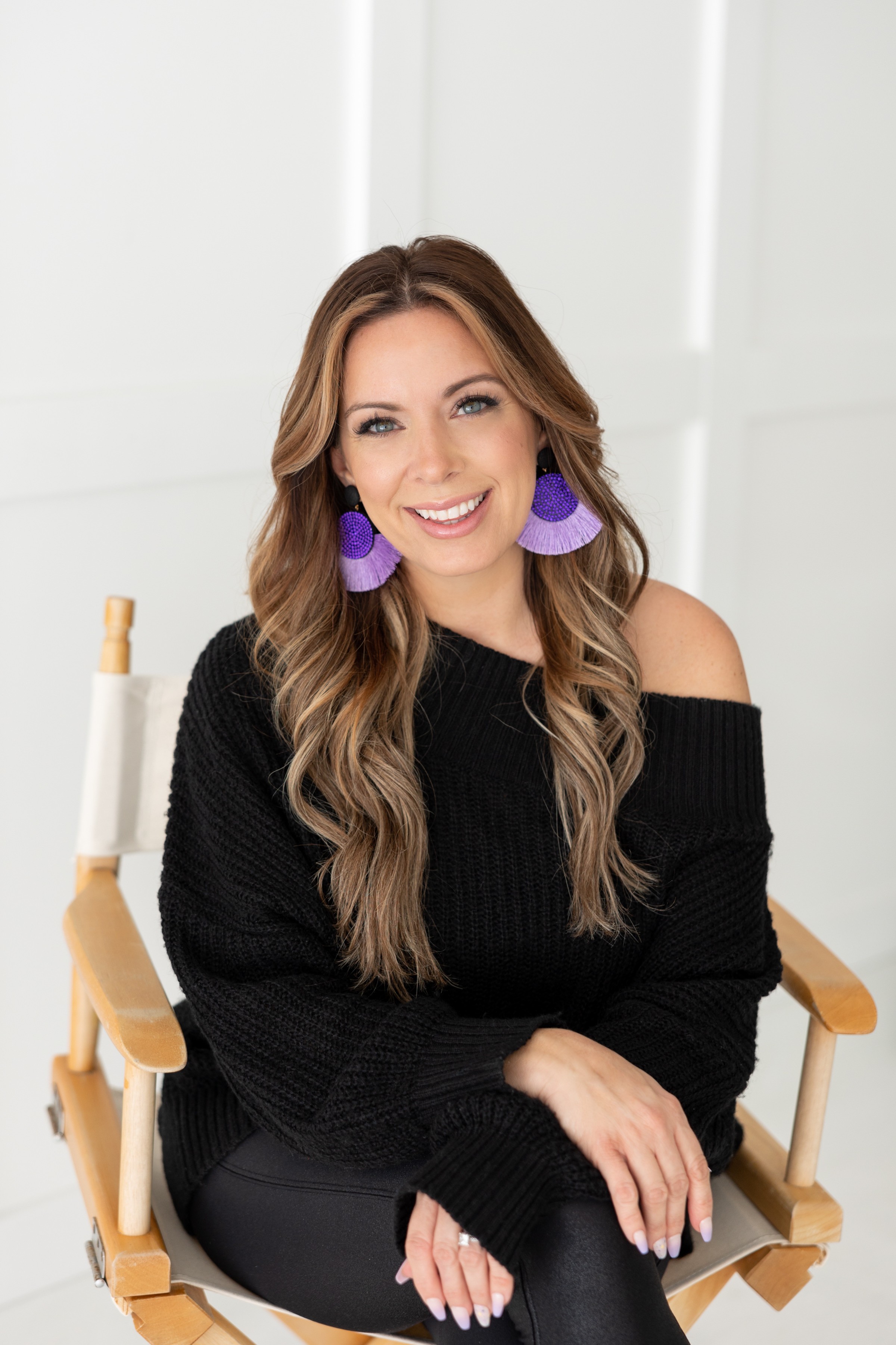 Smiling woman with long wavy hair wearing purple earrings and a black off-shoulder sweater, sitting on a wooden chair.