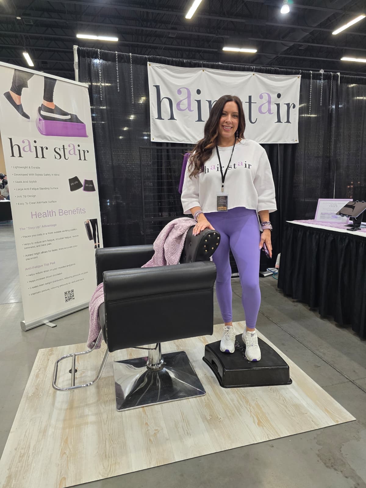 Woman standing on a platform at a hair and beauty event, smiling, with banners and tables in the background.