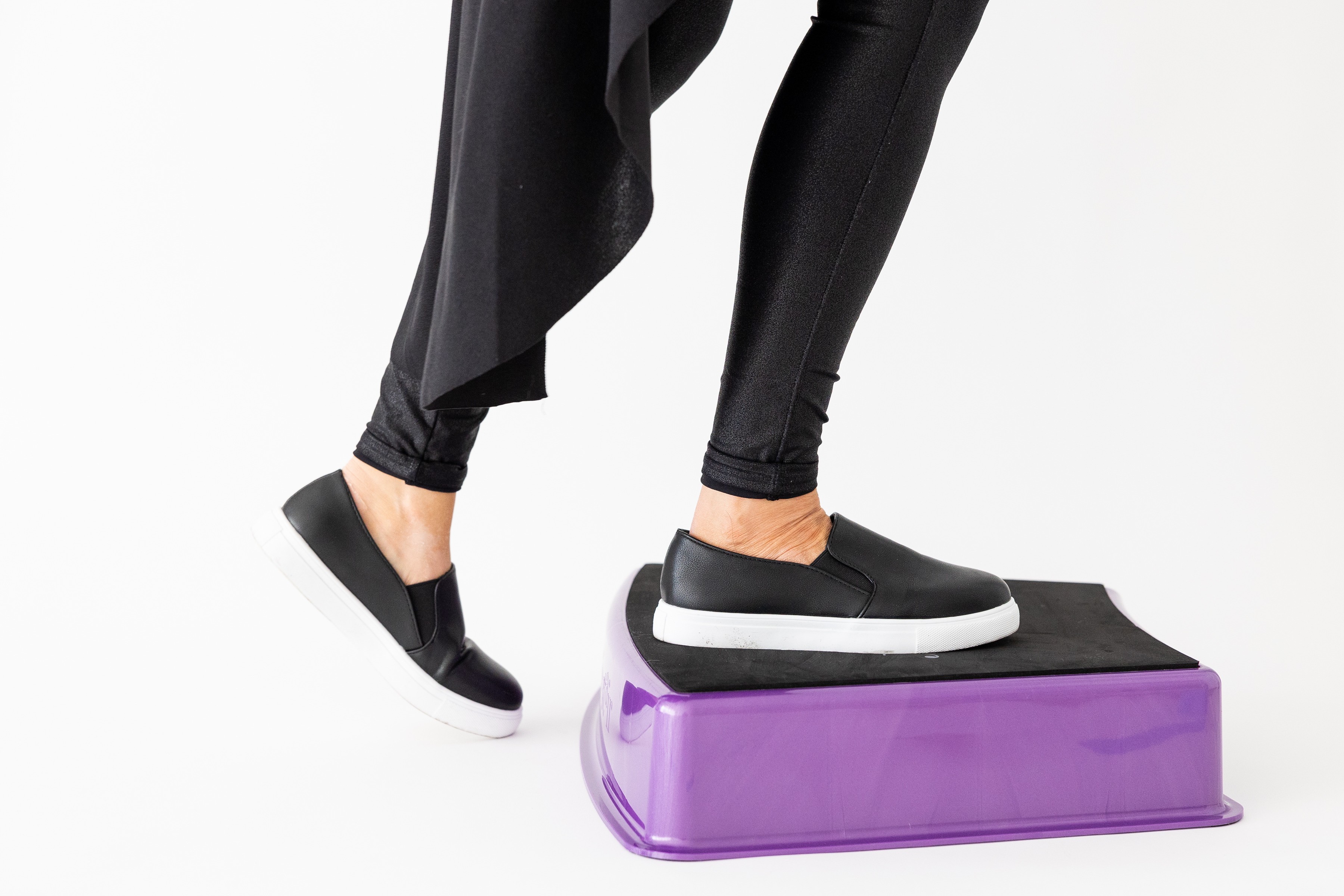 Person stepping onto a purple platform wearing black shoes and black pants, with another foot in the air. Plain white background.