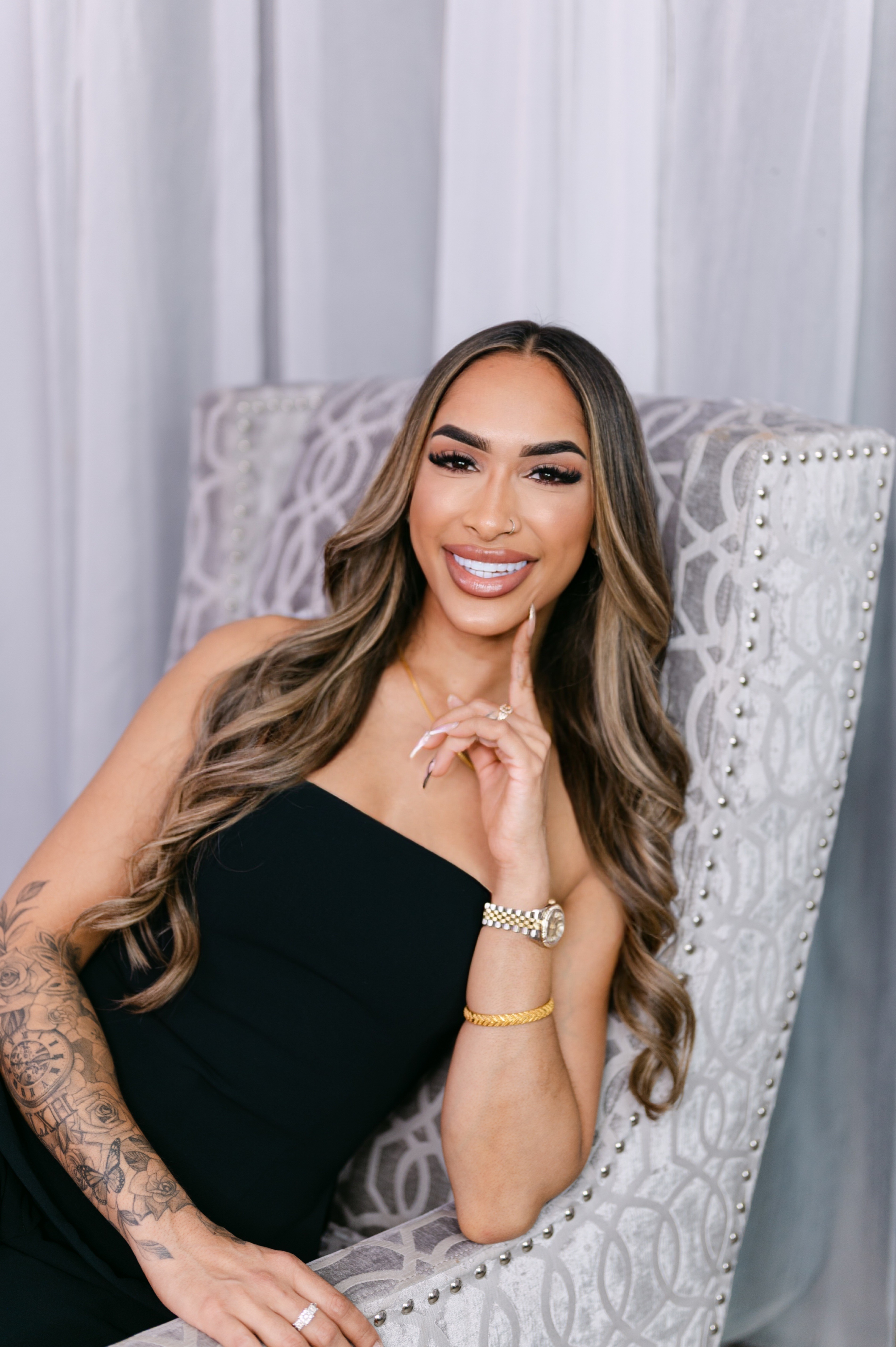 Smiling woman with long wavy hair, wearing a black dress, sitting on a patterned chair with a white curtain background.