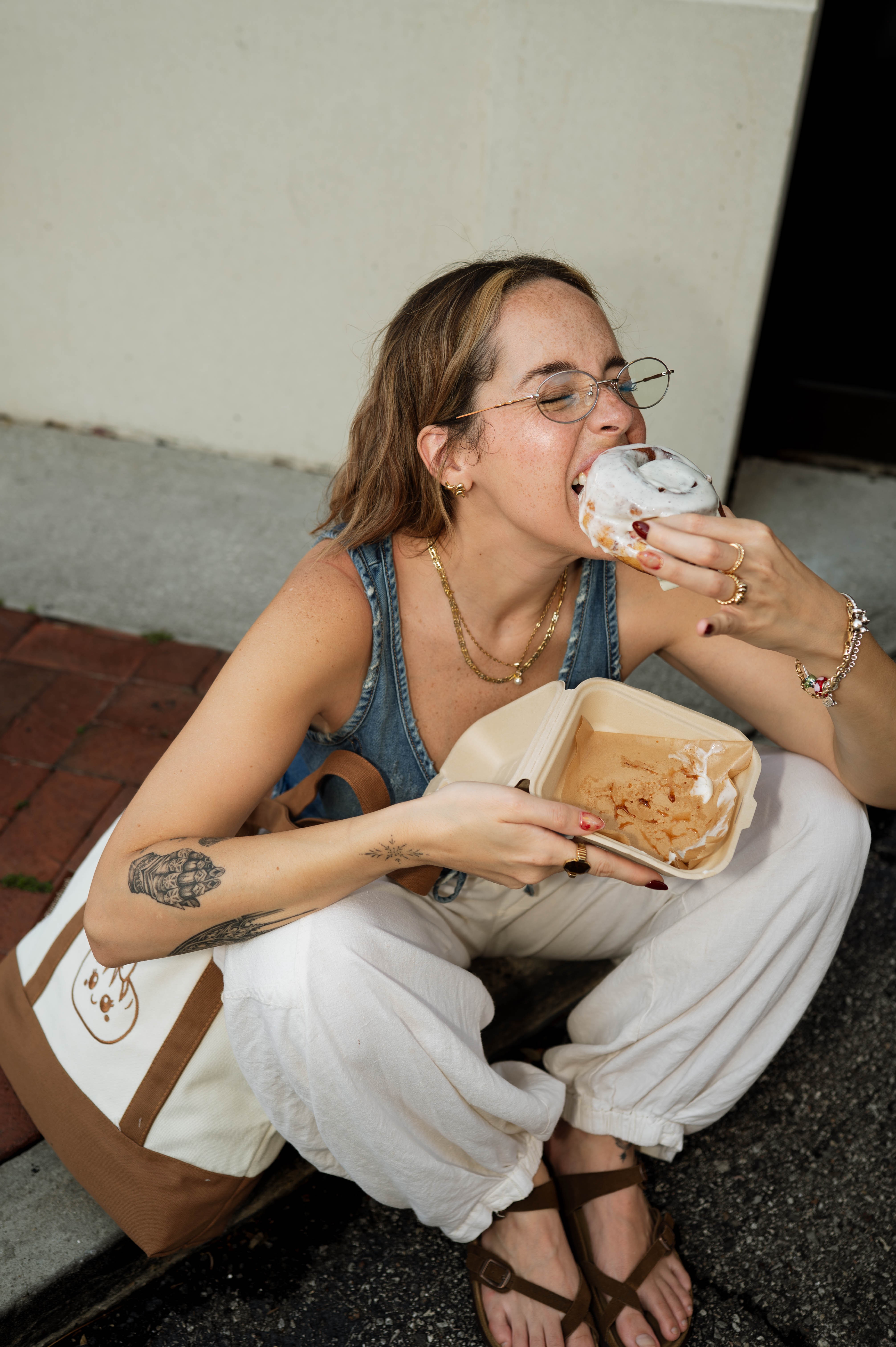 Woman with glasses and tattoos eating cake from a container, sitting on the ground with a bag nearby.