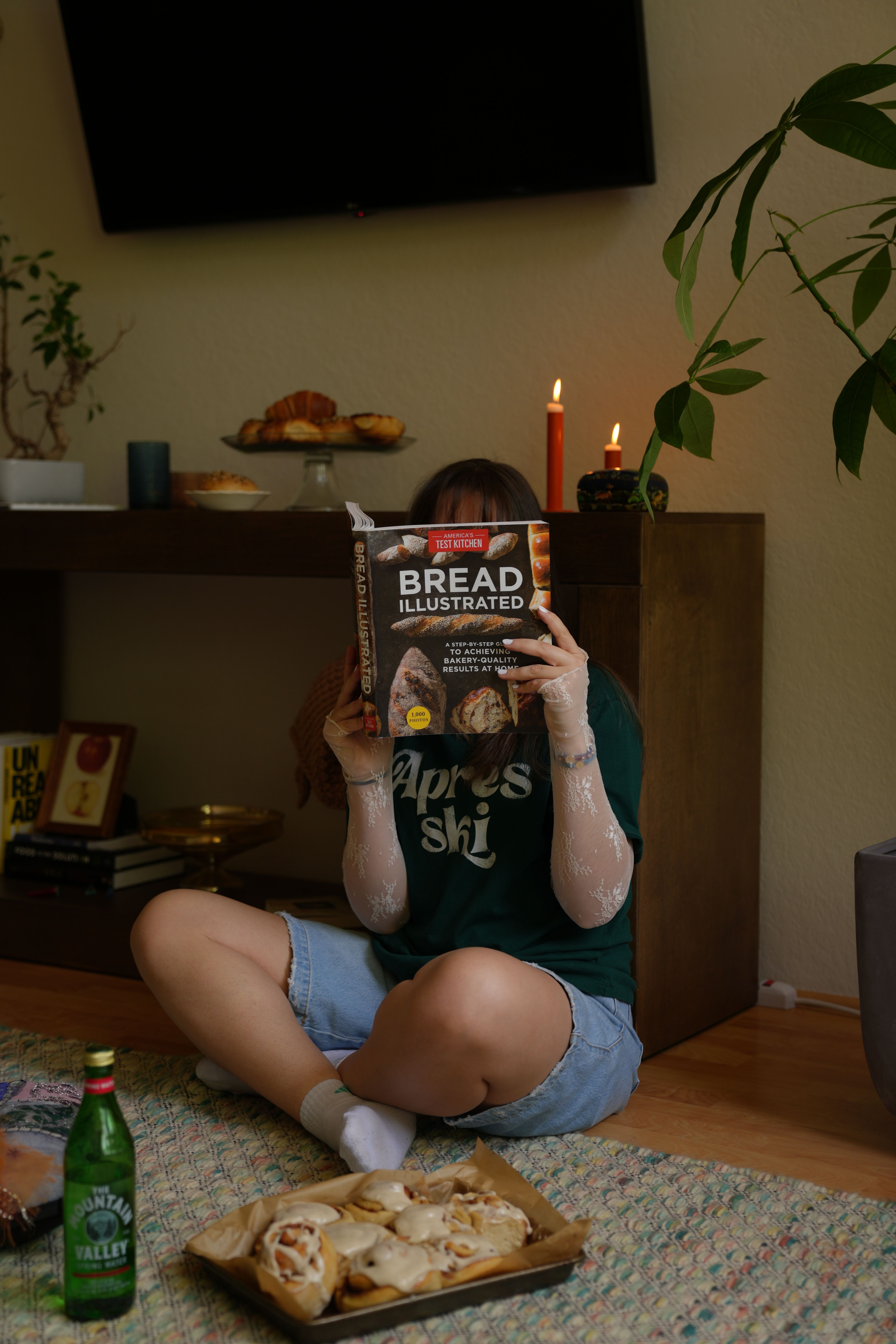Person sitting on the floor with food, wearing a black T-shirt and denim shorts, in a room with a TV and plant.
