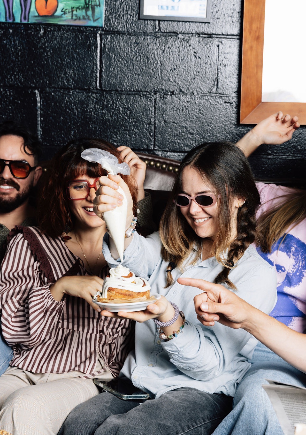 Group of four friends smiling, one holding a plate with a dessert, wearing sunglasses, in a cozy indoor setting.