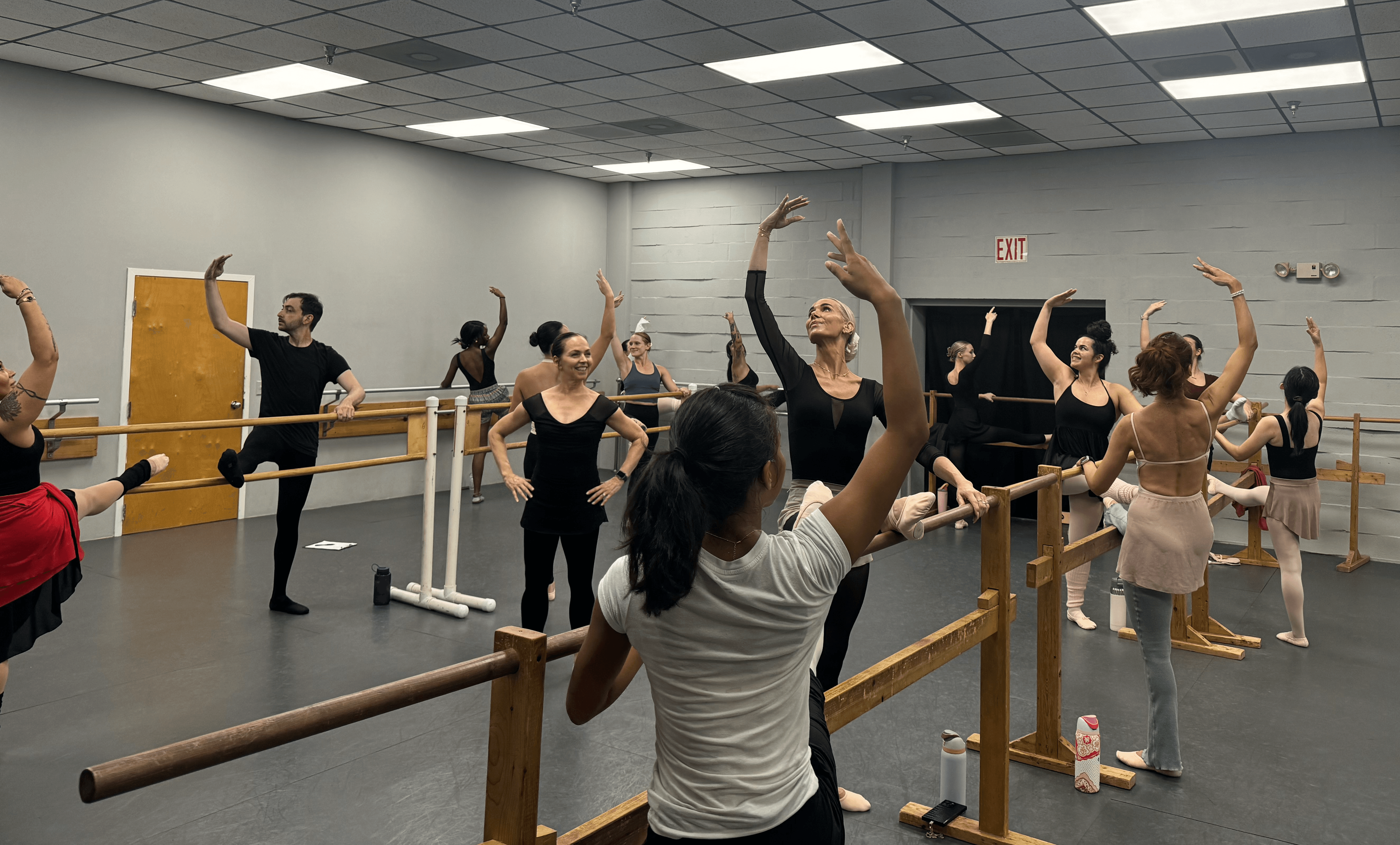 People in a dance studio practicing ballet, with some at the barre and others dancing in the center, all raising arms.
