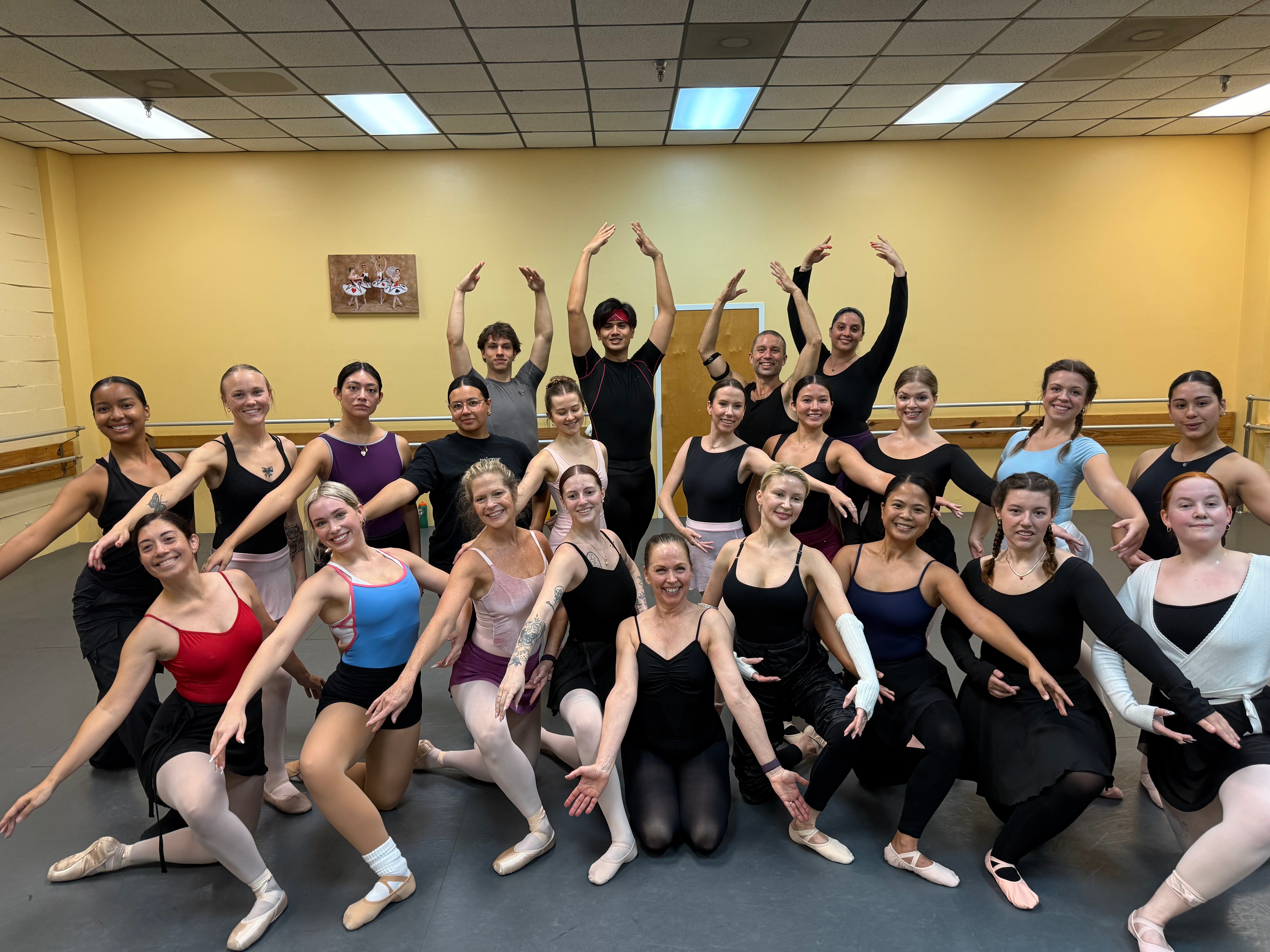 Group of ballet dancers in a dance studio, posing with arms raised and smiling, wearing leotards and tights.