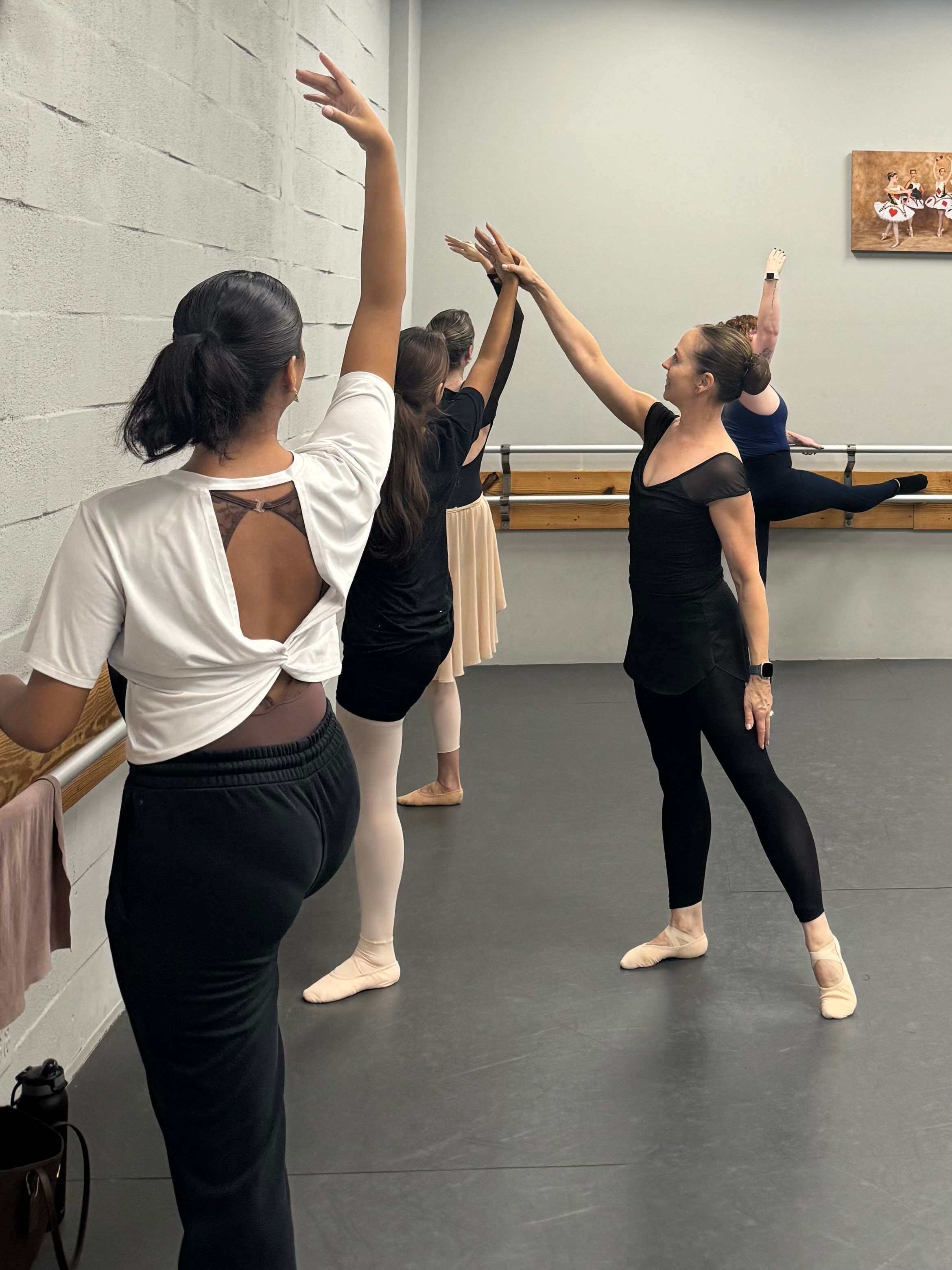 Four ballet dancers practicing at a barre in a dance studio, stretching with arms raised, wearing dance attire.