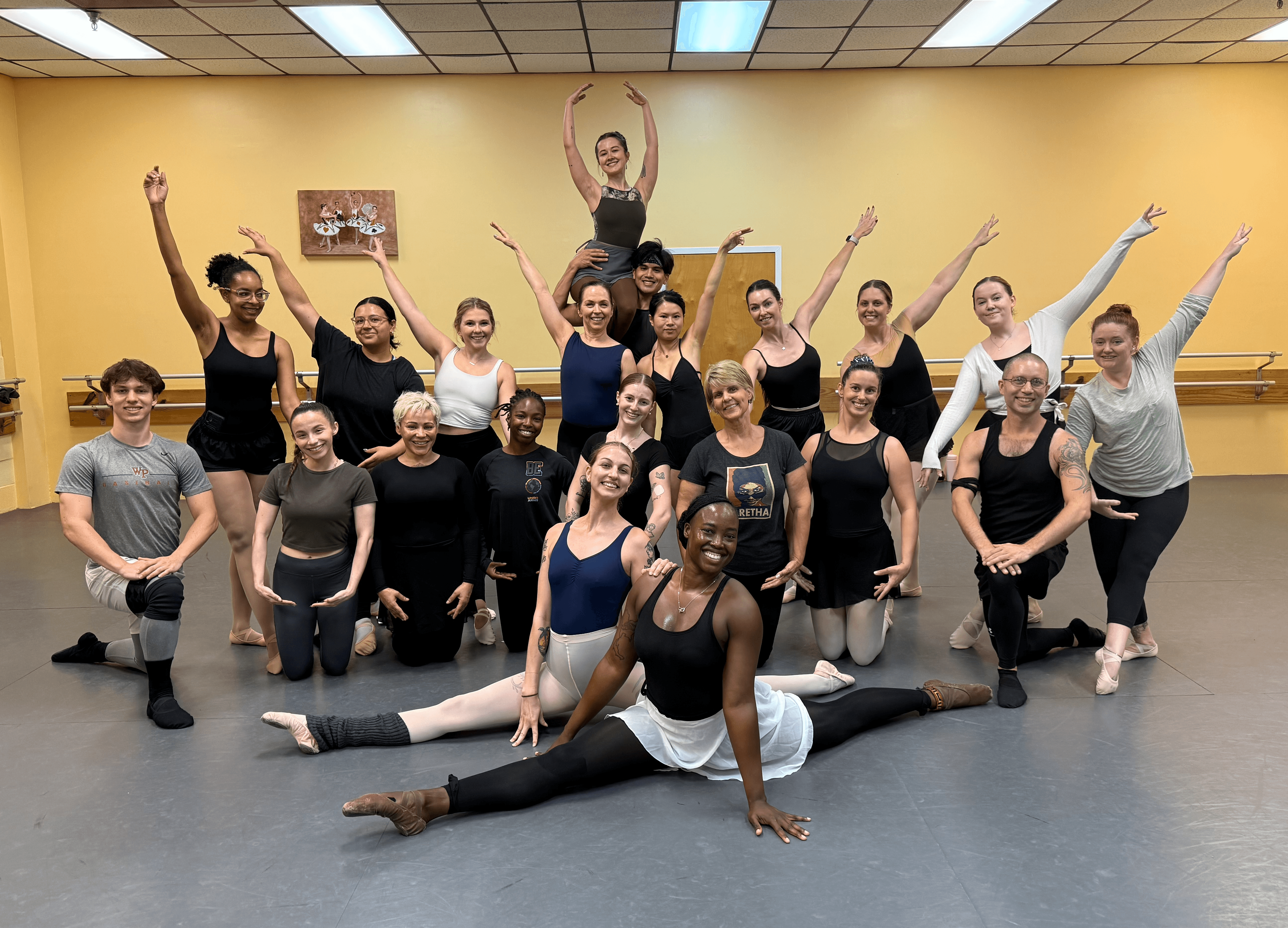 Group of dancers in a studio, some in splits, others standing with arms raised, smiling, yellow wall background.