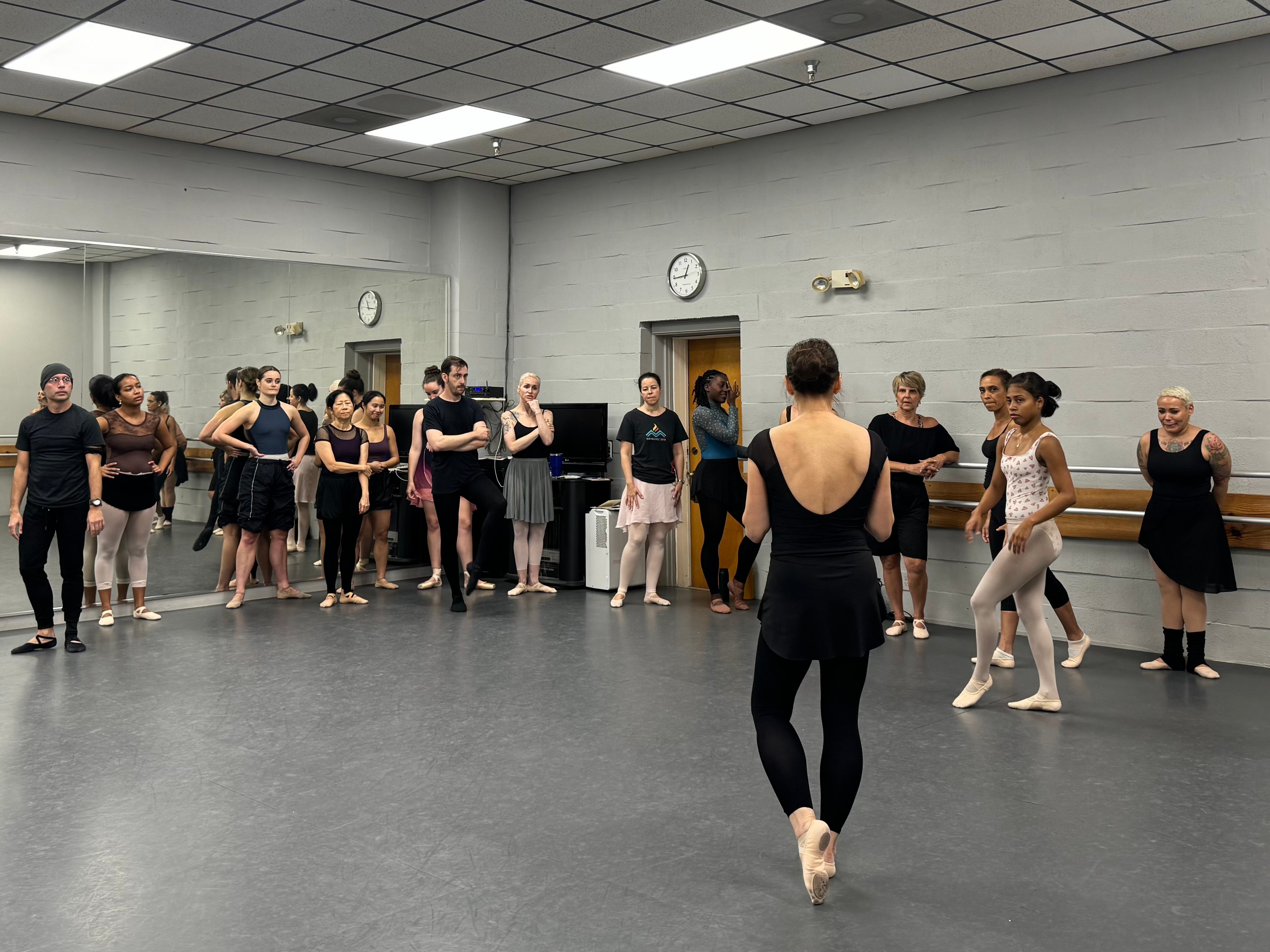 Ballet class with students standing in a circle, instructor in the center, in a dance studio with mirrors and clocks.