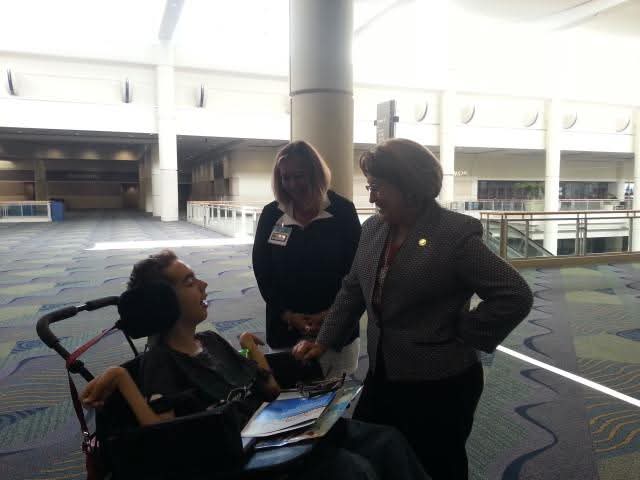 Three women engaged in conversation in a modern indoor space, one seated in a wheelchair, two standing, with books and papers.