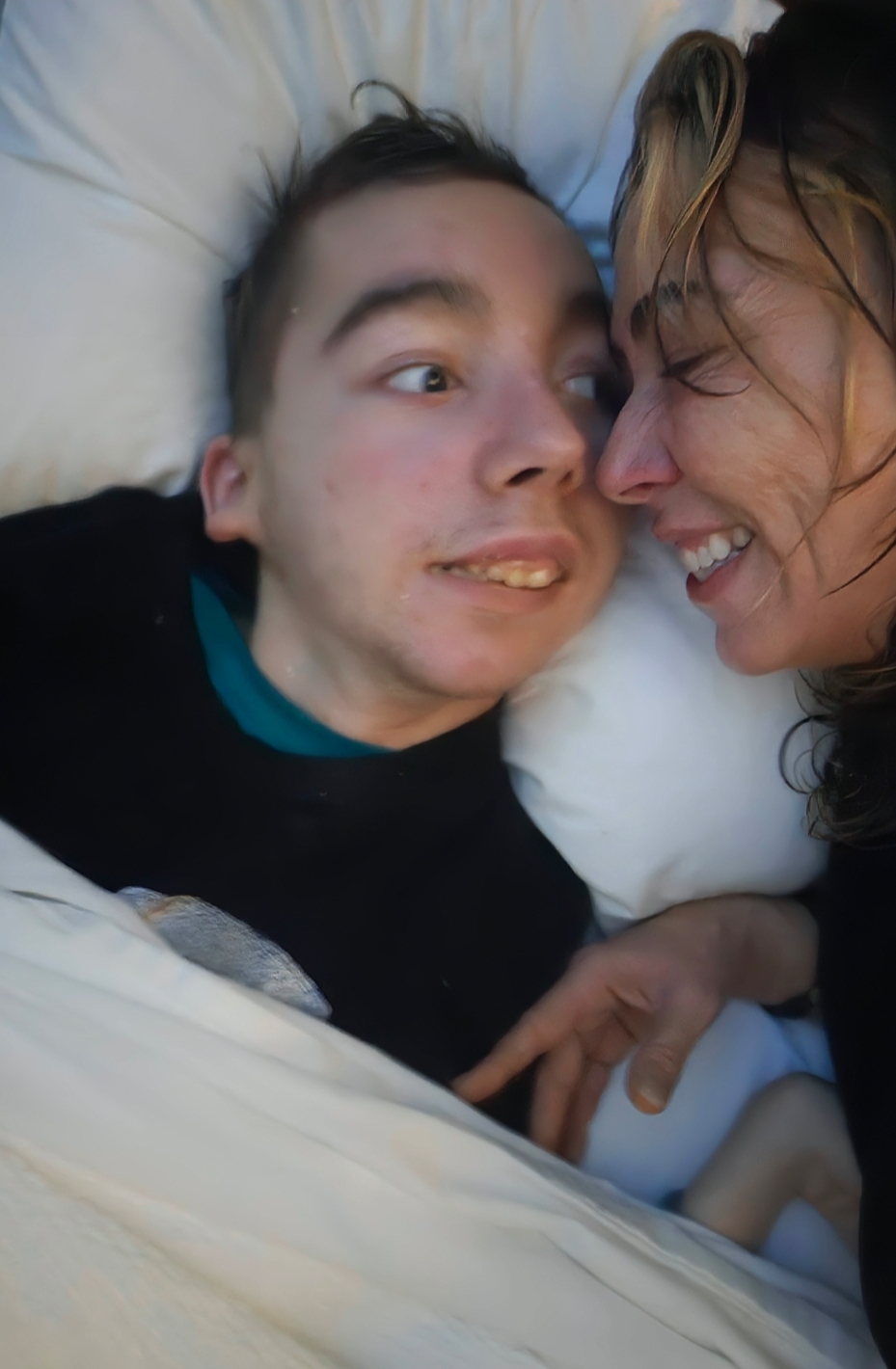 Two people lying close together, smiling and touching faces, on a bed with white sheets.