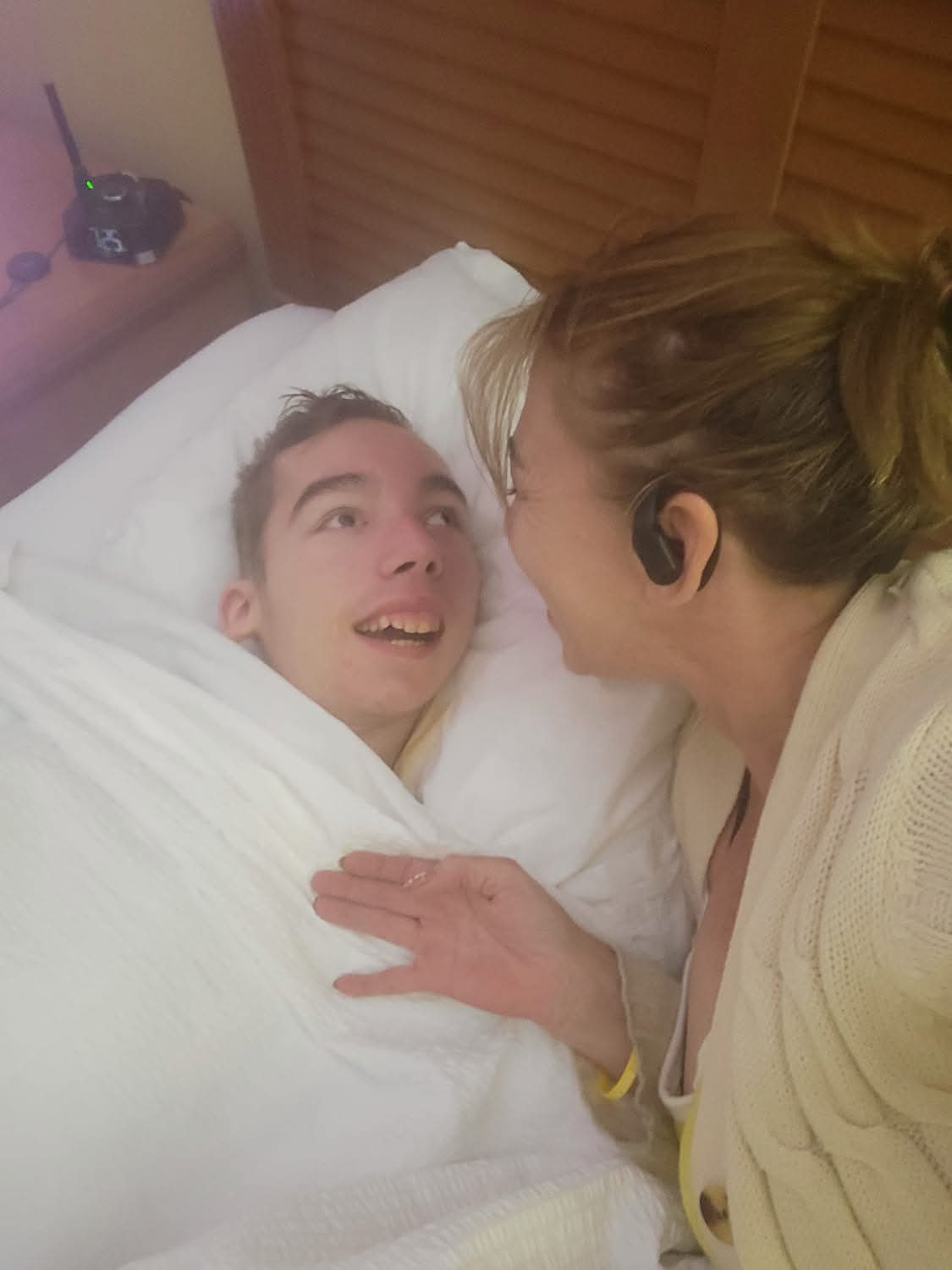 A woman leans over a man in bed, both smiling and engaging closely, with a wooden headboard in the background.