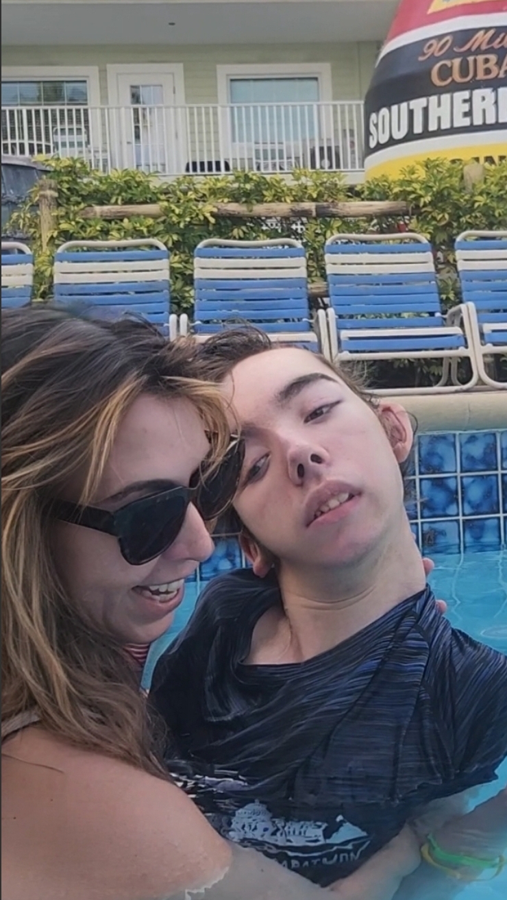 Two people in a swimming pool, one wearing sunglasses, close-up selfie with pool chairs and a sign in the background.