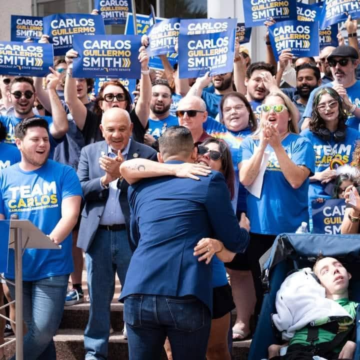 Crowd of people at a rally holding signs supporting Carlos Guillermo Smith, with two men hugging in the center.
