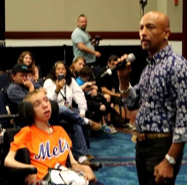 Man holding microphone speaking to audience in a room with seated people, some taking photos, young girl in orange jersey in foreground.