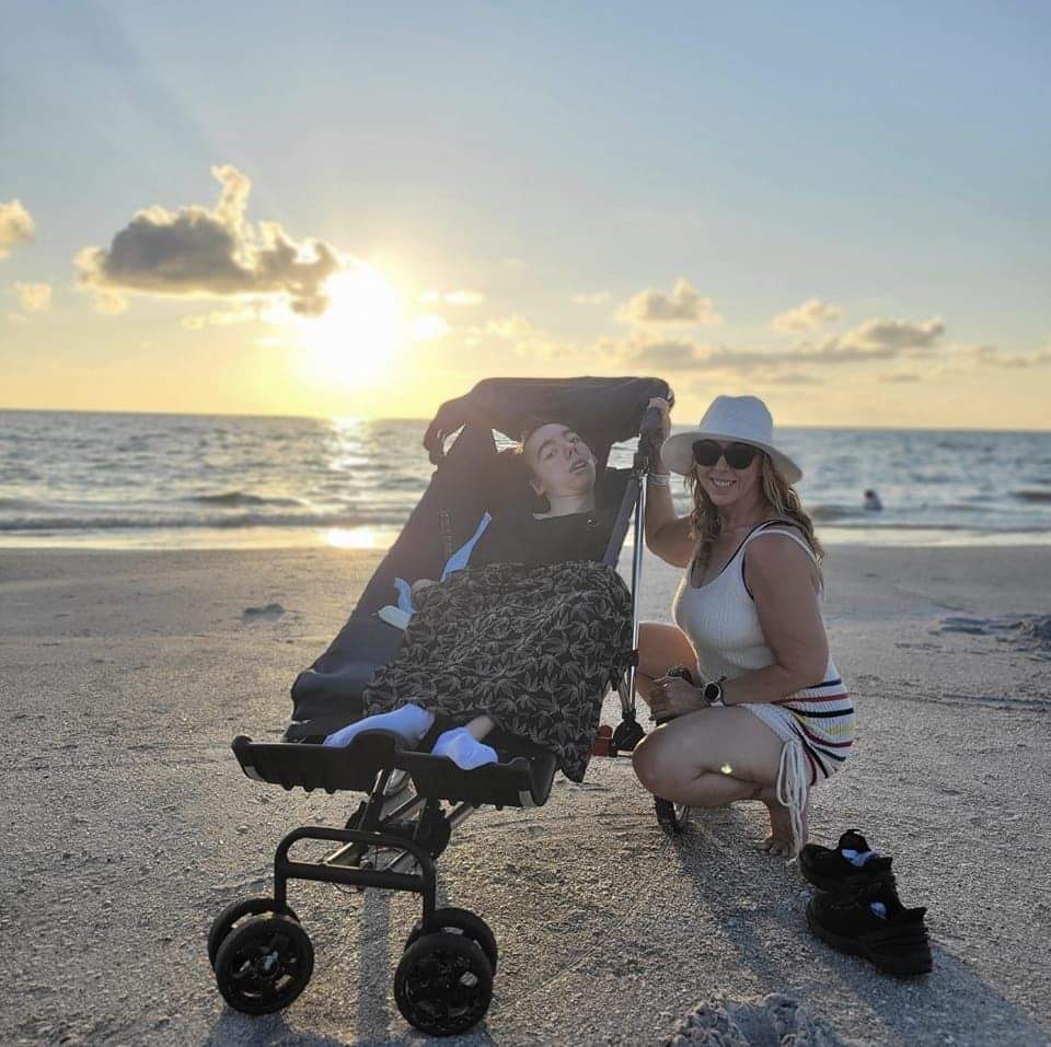 Two women with a stroller on a beach at sunset, one sitting and one squatting, both smiling, wearing sunglasses and hats.
