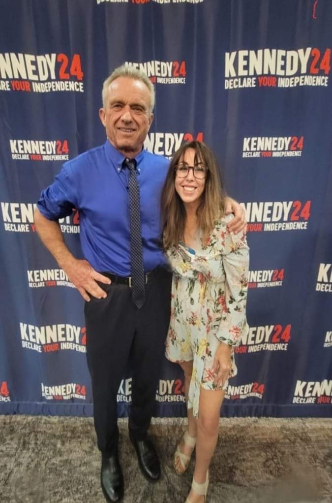 A man and a woman stand together in front of a backdrop with 'Kennedy 24' logos, smiling at the camera.