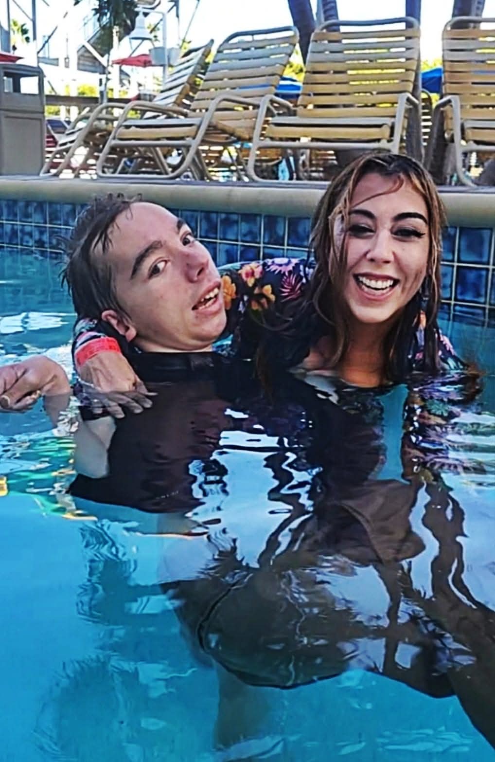 Two women smiling and enjoying a swim in a pool, with pool chairs and umbrellas in the background.