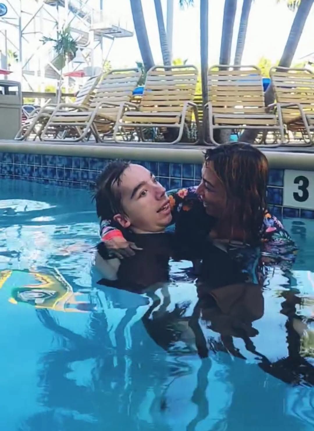 Two women in a swimming pool, one holding the other, with pool chairs and umbrellas in the background.